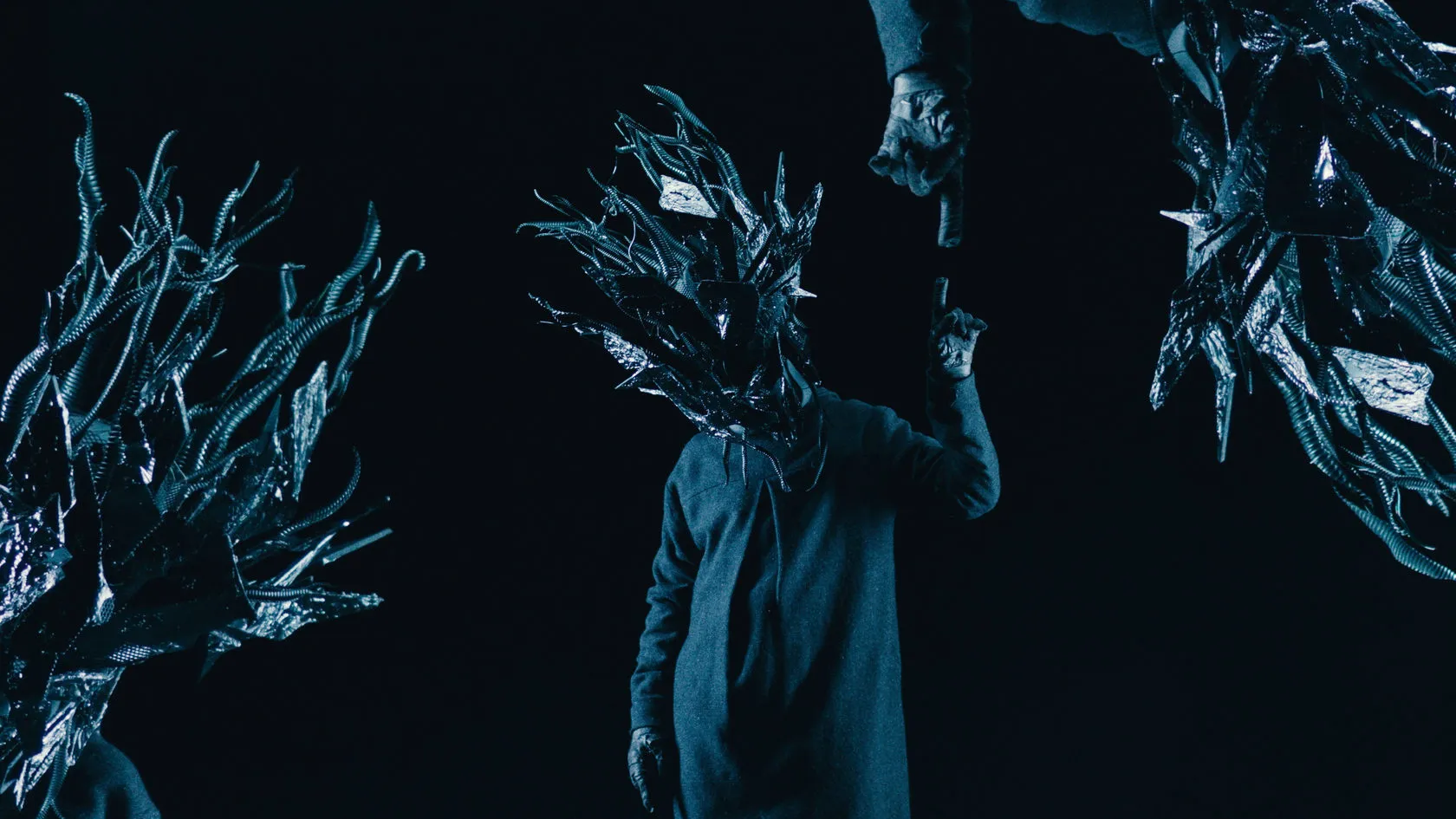 Figures in spiky black masks under blue light; one points up as a gloved hand reaches down to him.
