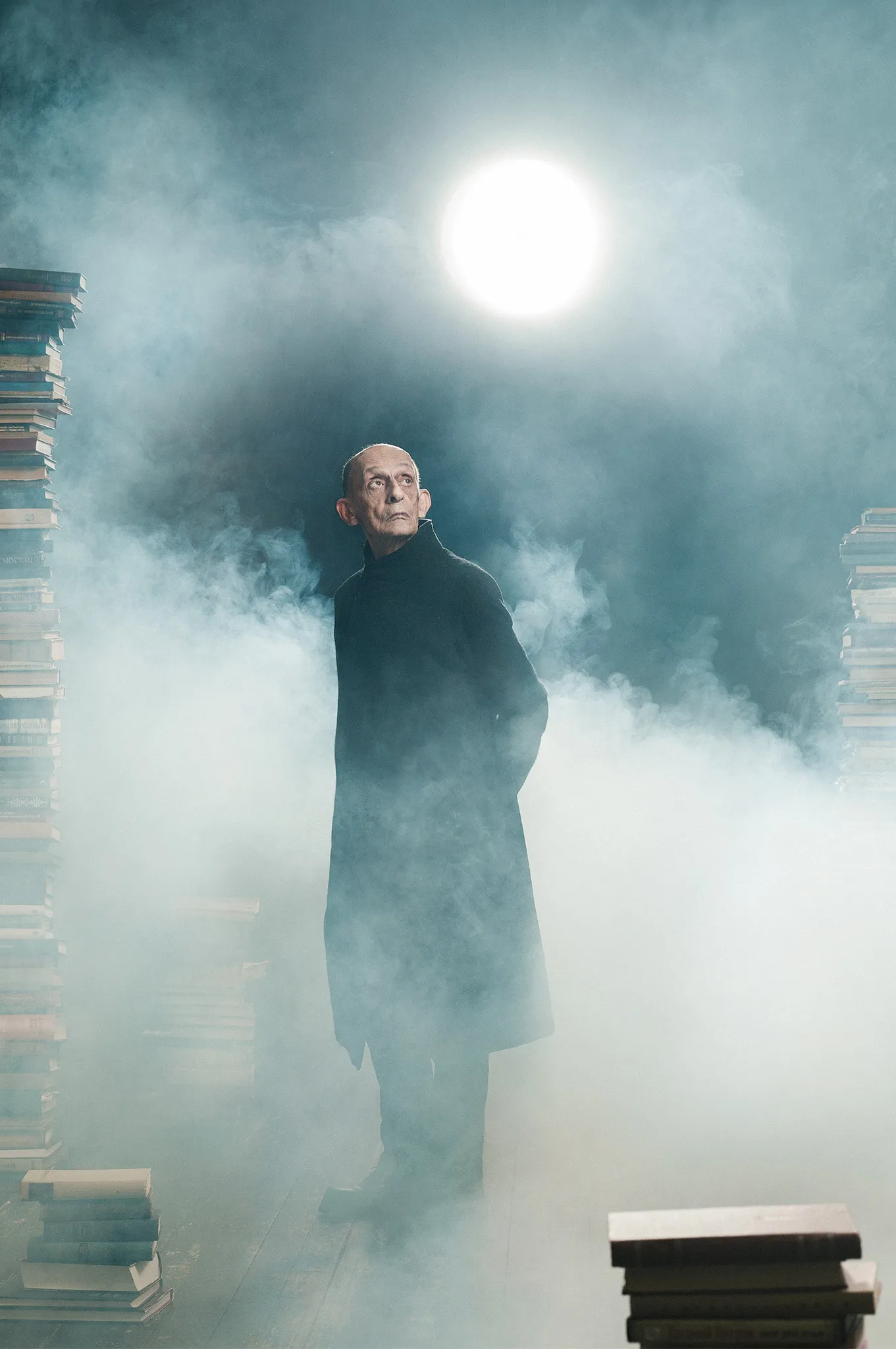 An old man in a long dark coat stands in thick fog under a bright light, between stacks of books.