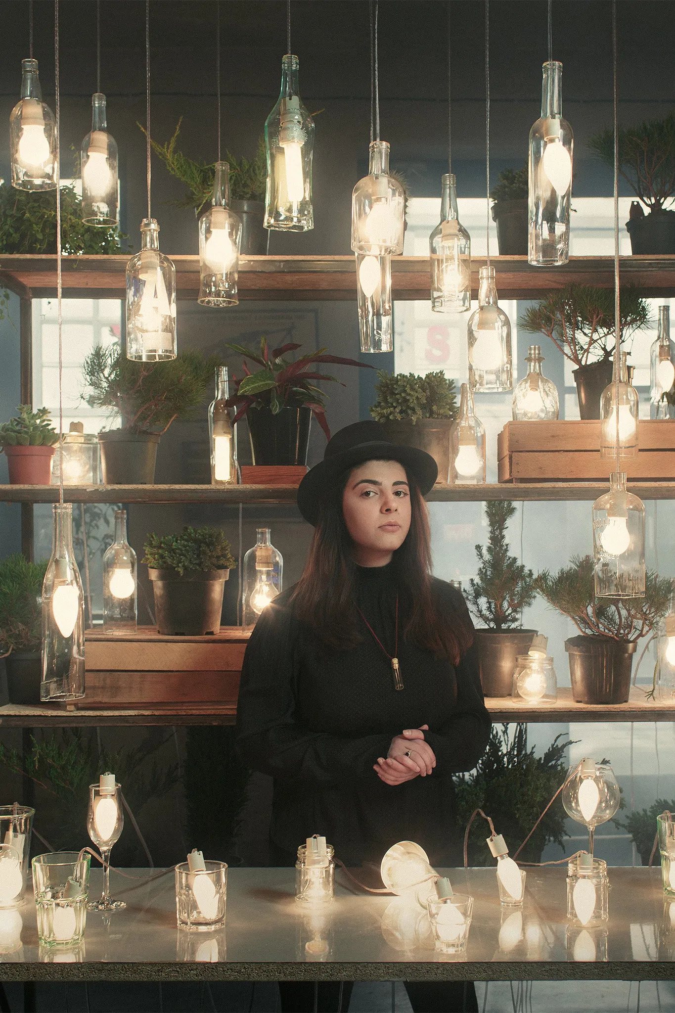 A woman in a black hat stands behind a table of glowing lamps and bottles, with plants on shelves behind her.
