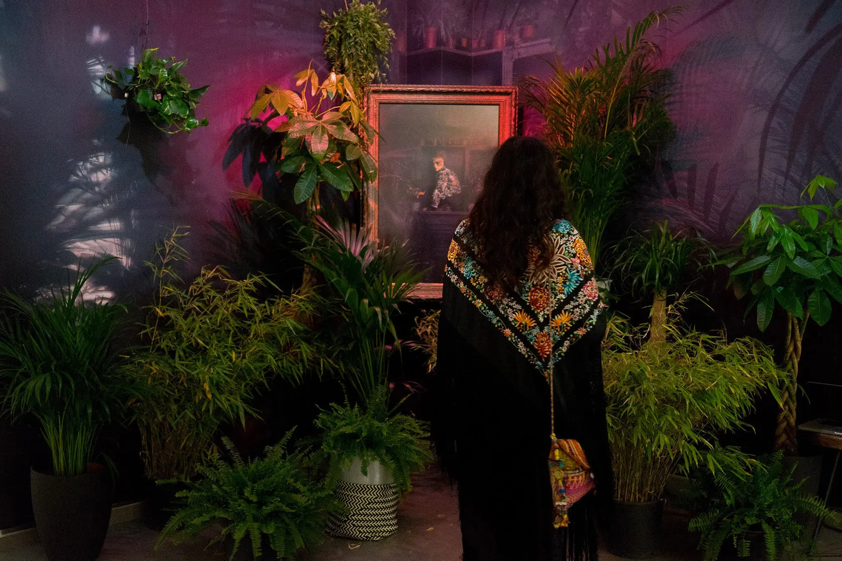 A woman in an embroidered shawl looks at a framed portrait hung among potted plants at an exhibition.