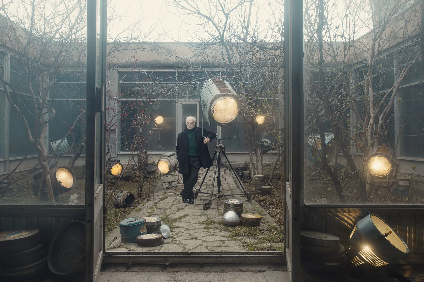 An elderly man stands among old studio spotlights in a misty courtyard, seen through open glass doors.