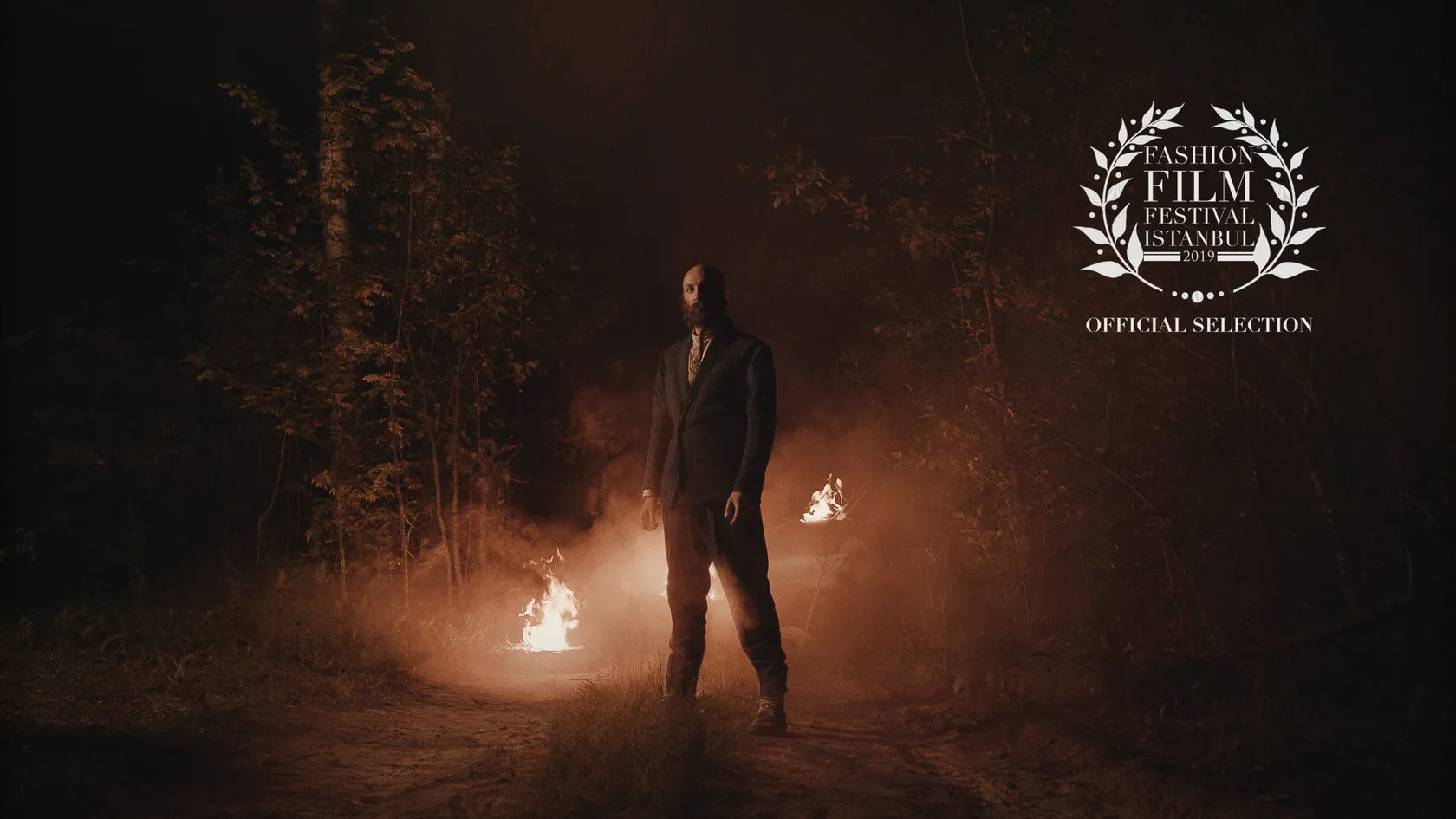 A man in a suit stands between small fires in a dark forest, beside a Fashion Film Festival Istanbul selection laurel.