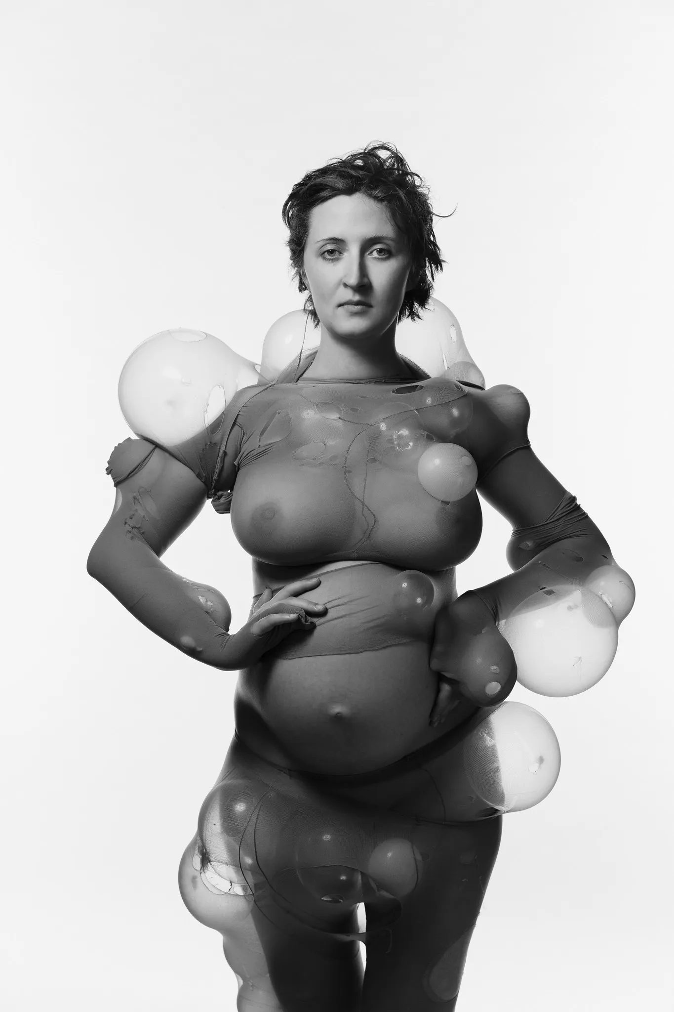 A pregnant woman wears torn sheer tights stretched over balloons, in black and white.