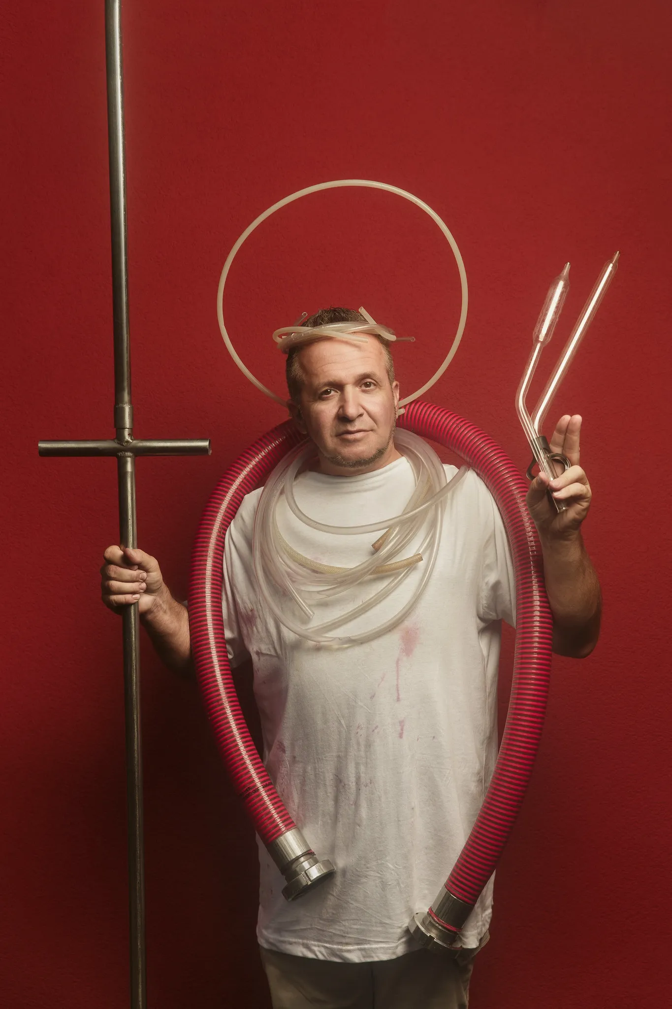 A man in a wine-stained T-shirt poses like a saint, with a tubing halo, a red hose, a steel pole and glass pipettes.