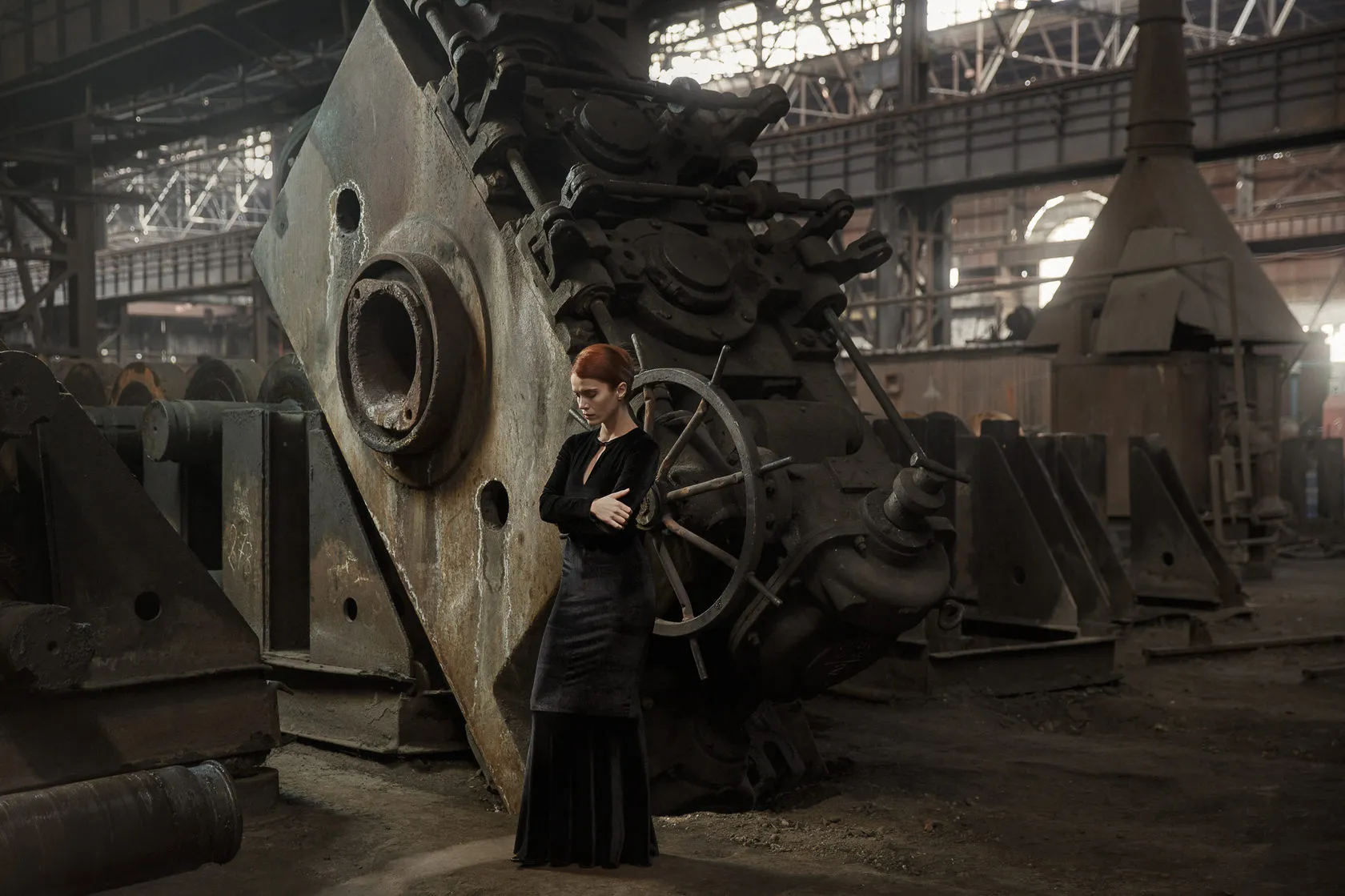 A woman in a long black dress stands beside giant rusted machinery in a vast factory hall.