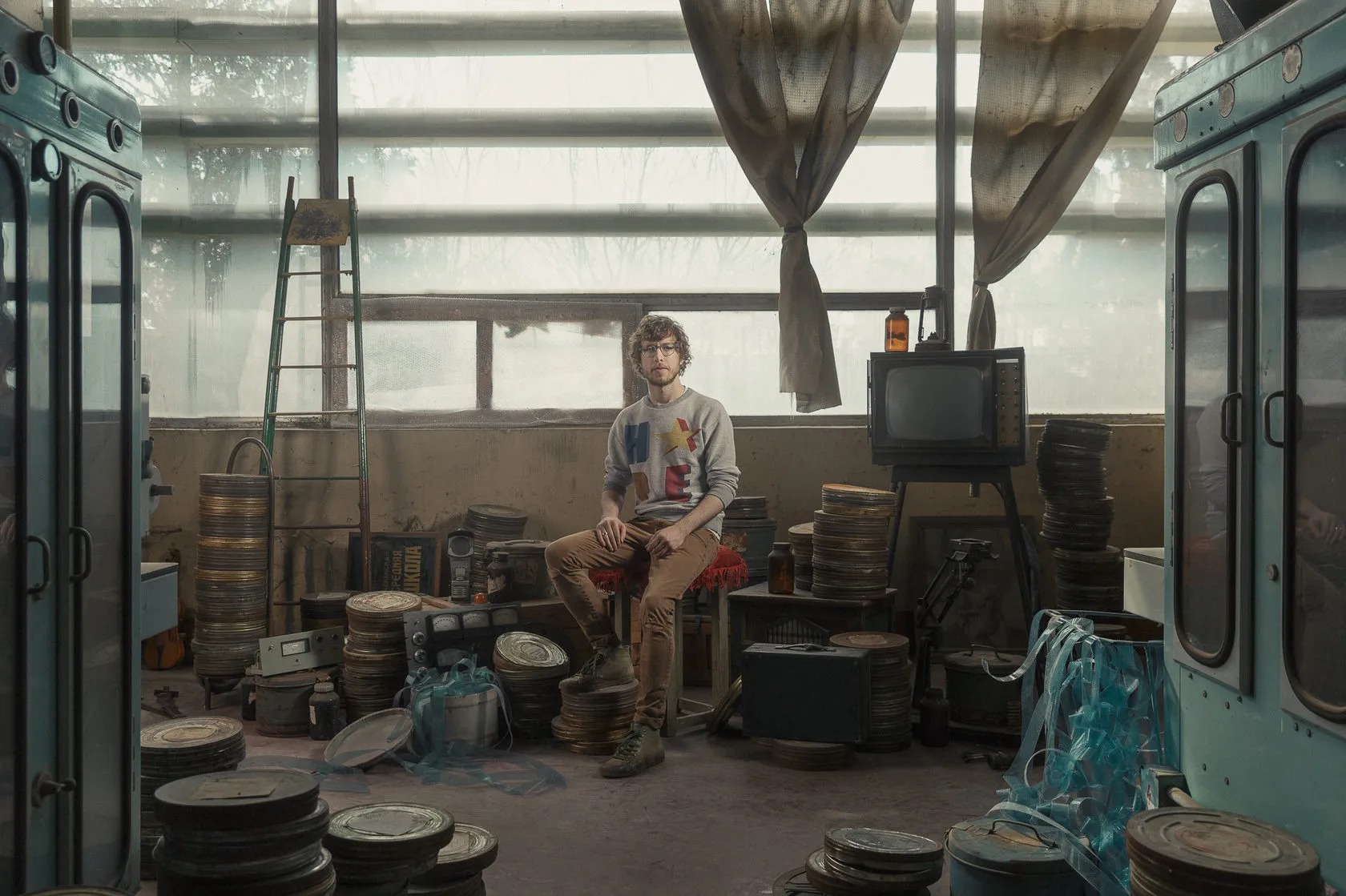 A man in glasses sits on a stool in a projection room full of film cans, an old TV and tangled film.