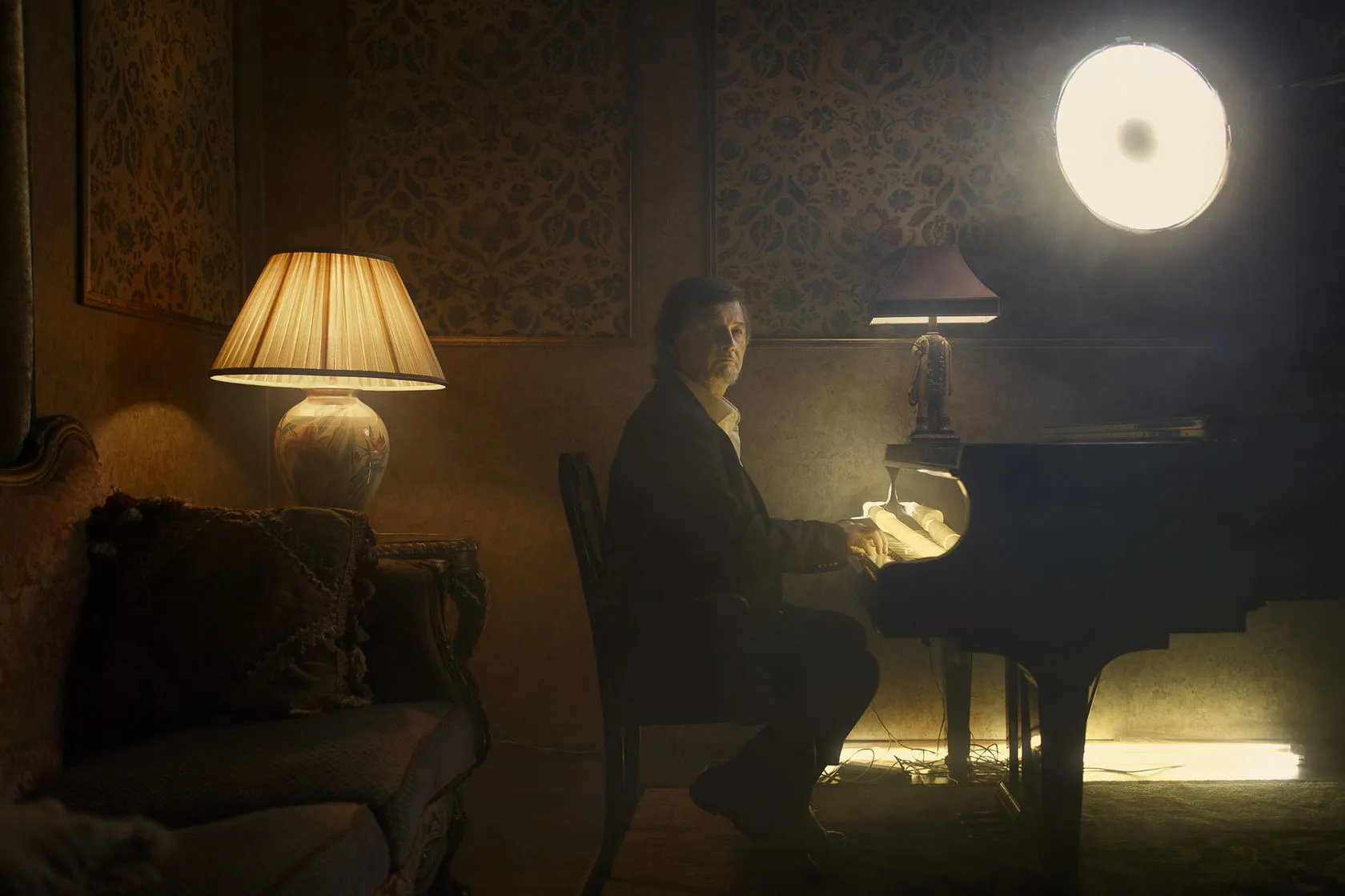 Alexey Rybnikov plays a grand piano in a dim room lit by a lamp and a round light.