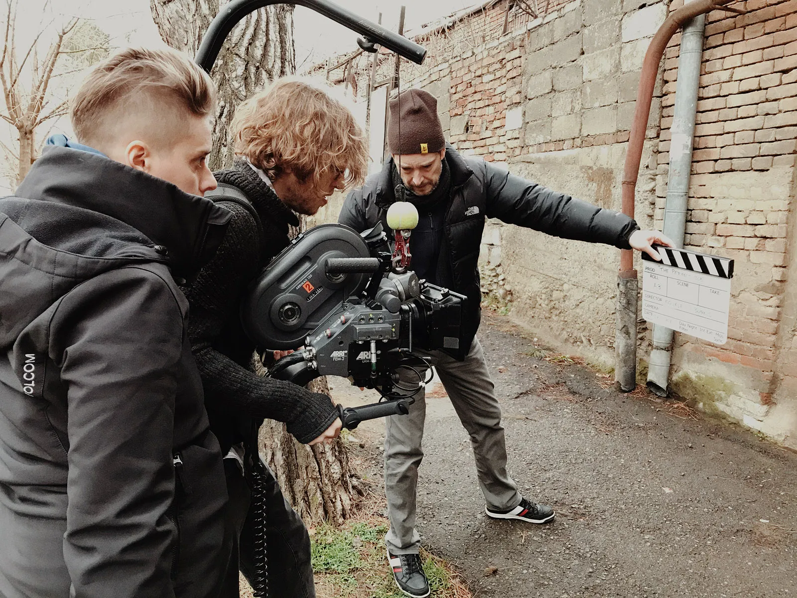 Three crew members at a 35mm film camera, one holding out a clapperboard, by a brick wall.