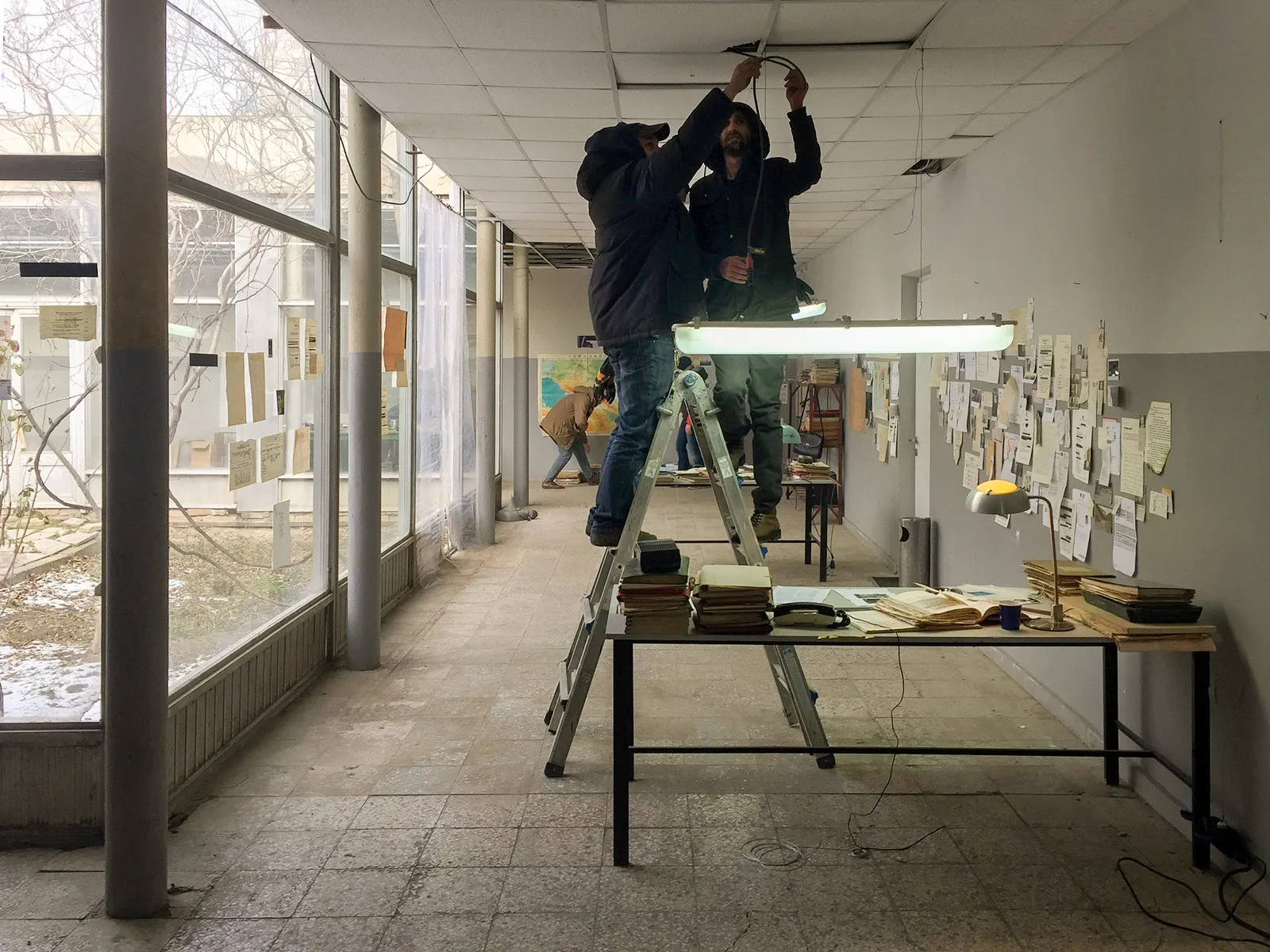Two men on a stepladder hang a fluorescent lamp over a desk in the hall.