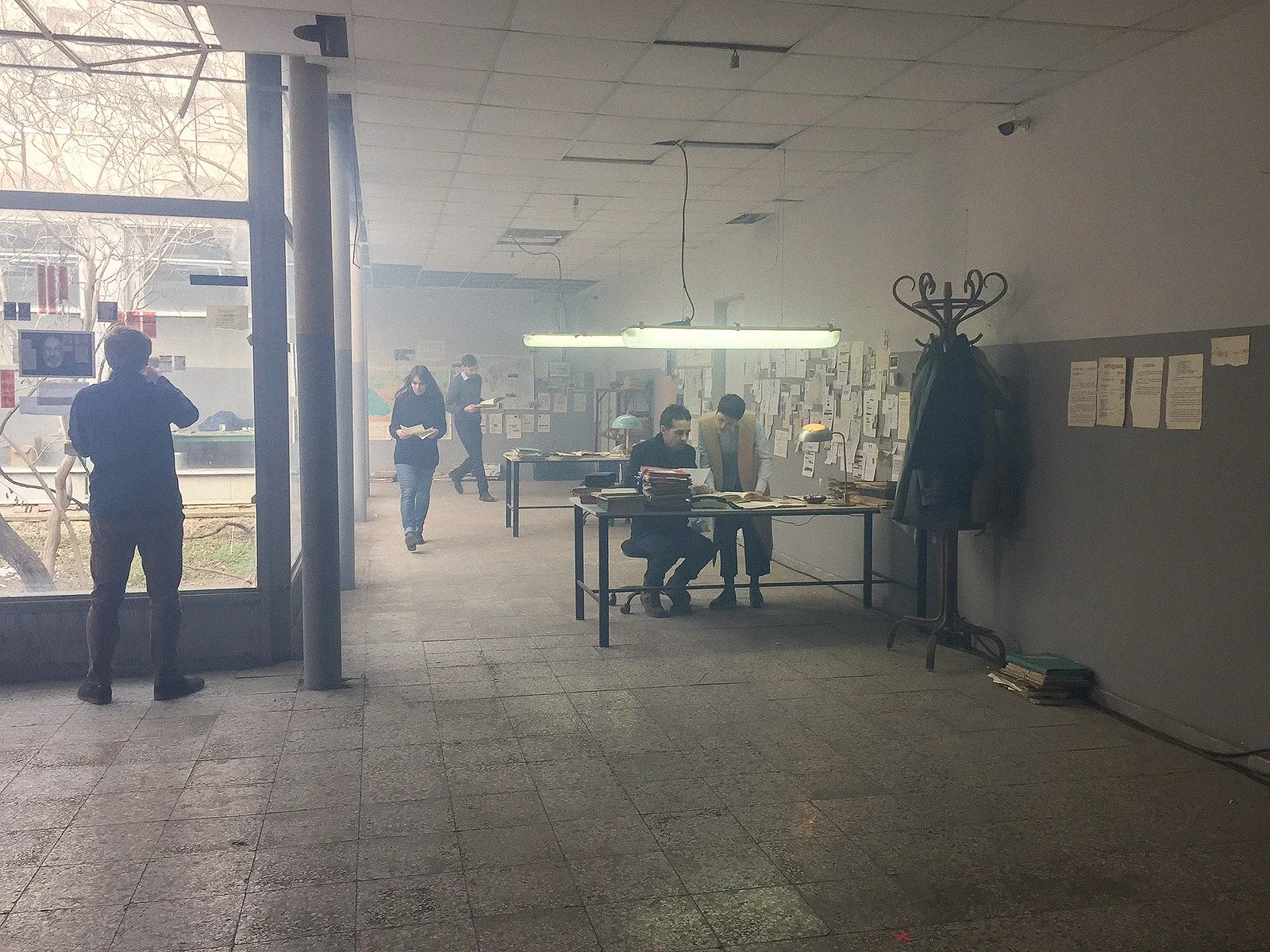 Actors at a desk under fluorescent lamps in the hazy hall, a man by the window.