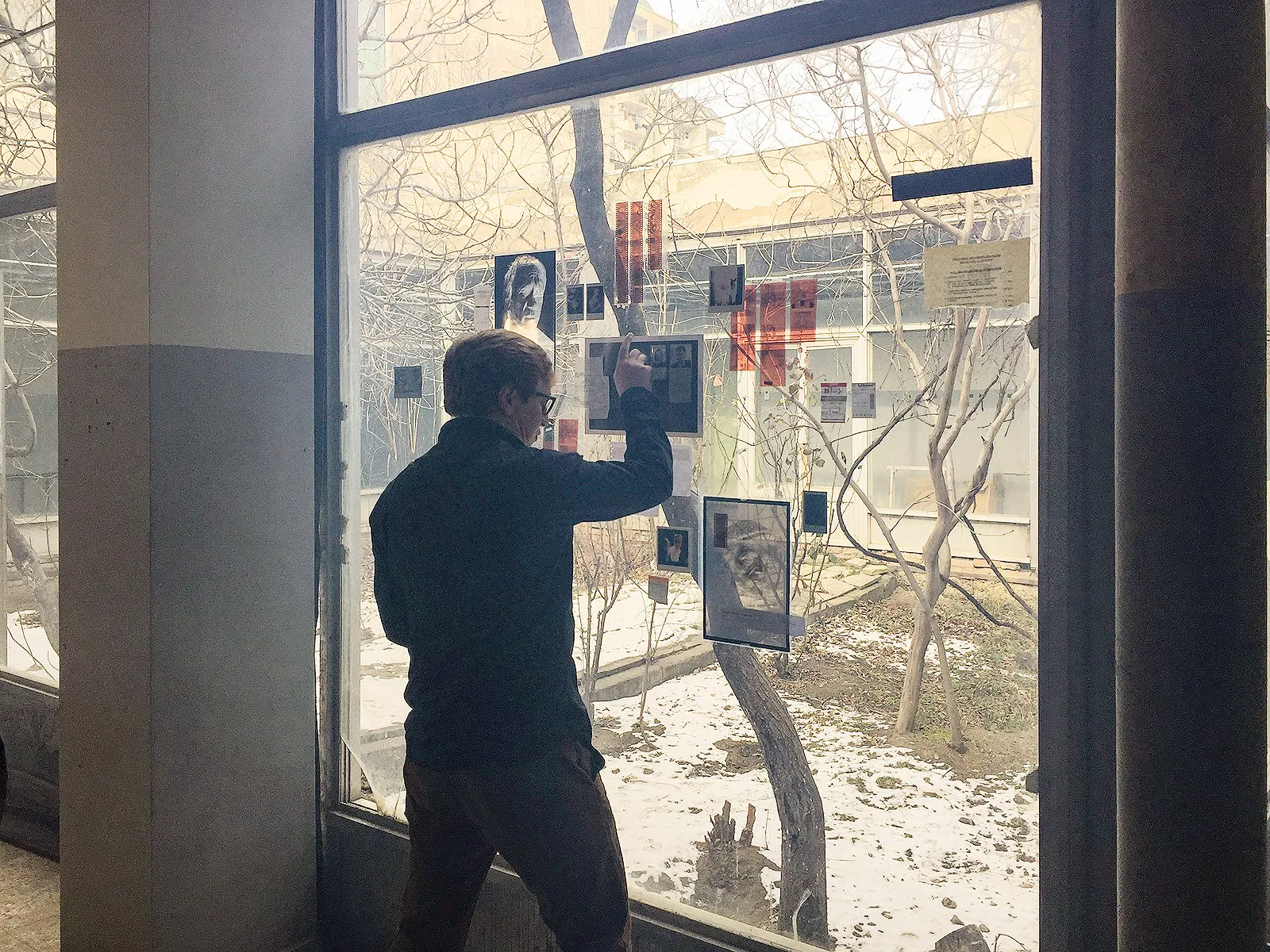 A man pins photos and papers to a big window looking onto a snowy courtyard.