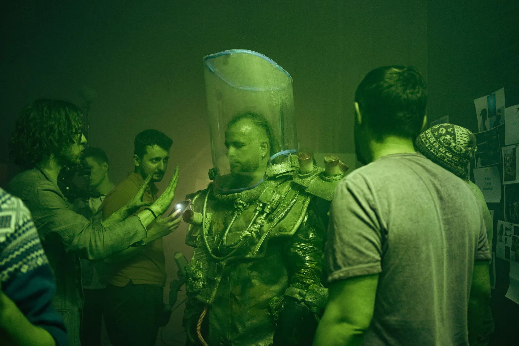 Buritō Kōsu by Andrei Kovalev: students fit an oversized clear helmet onto a model during a workshop, lit green.