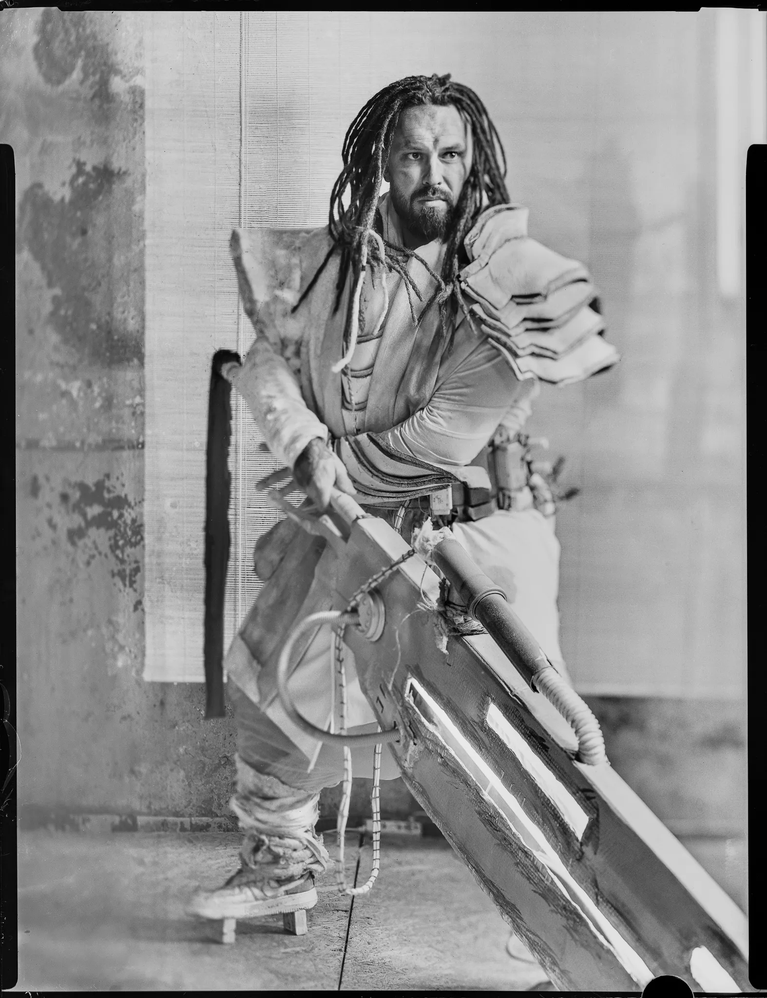 A man with dreadlocks in padded armour swings a huge blade, on black-and-white sheet film.