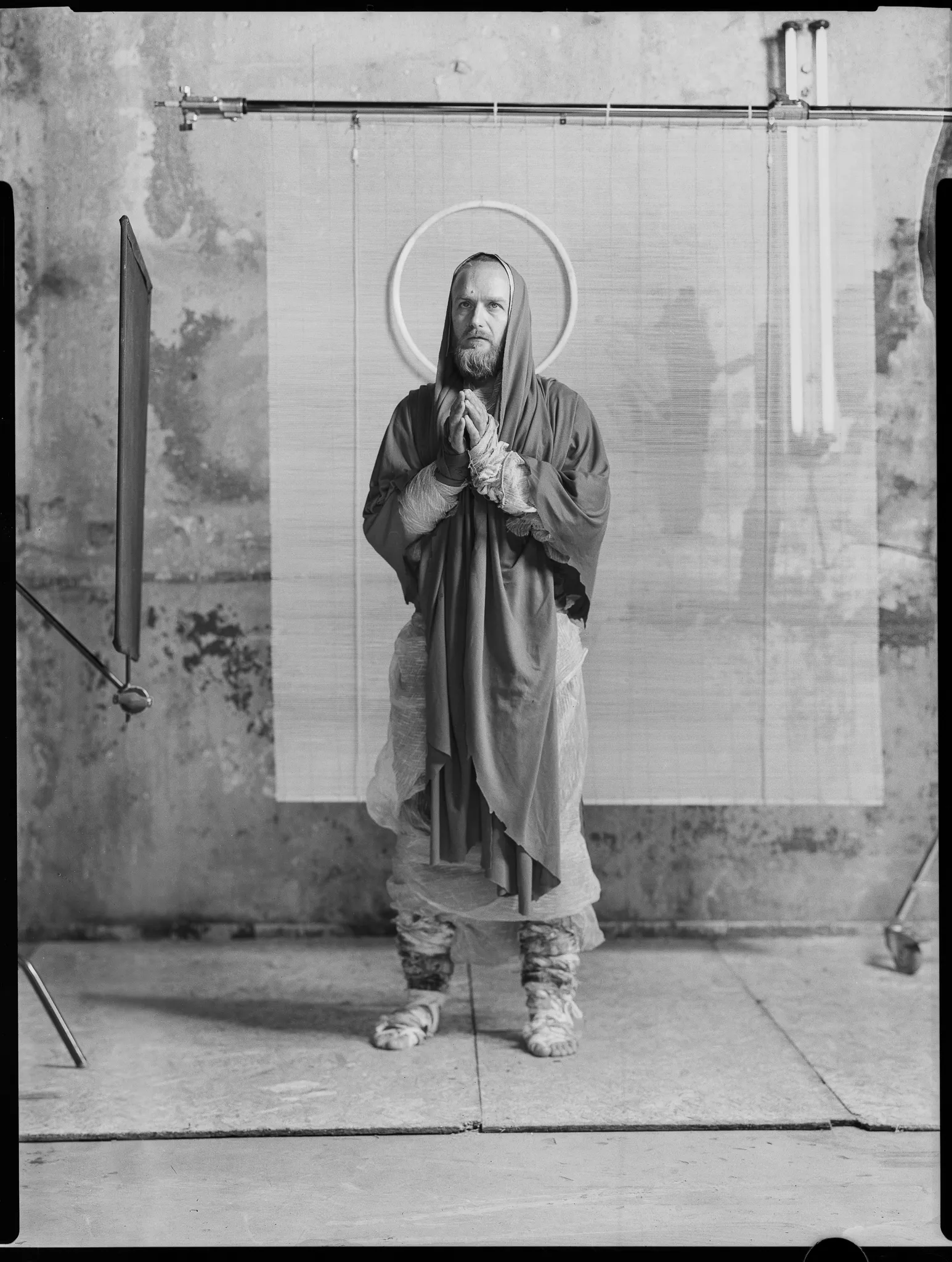 The Cyber Monk in a hooded robe and halo stands with palms pressed together, on black-and-white sheet film.