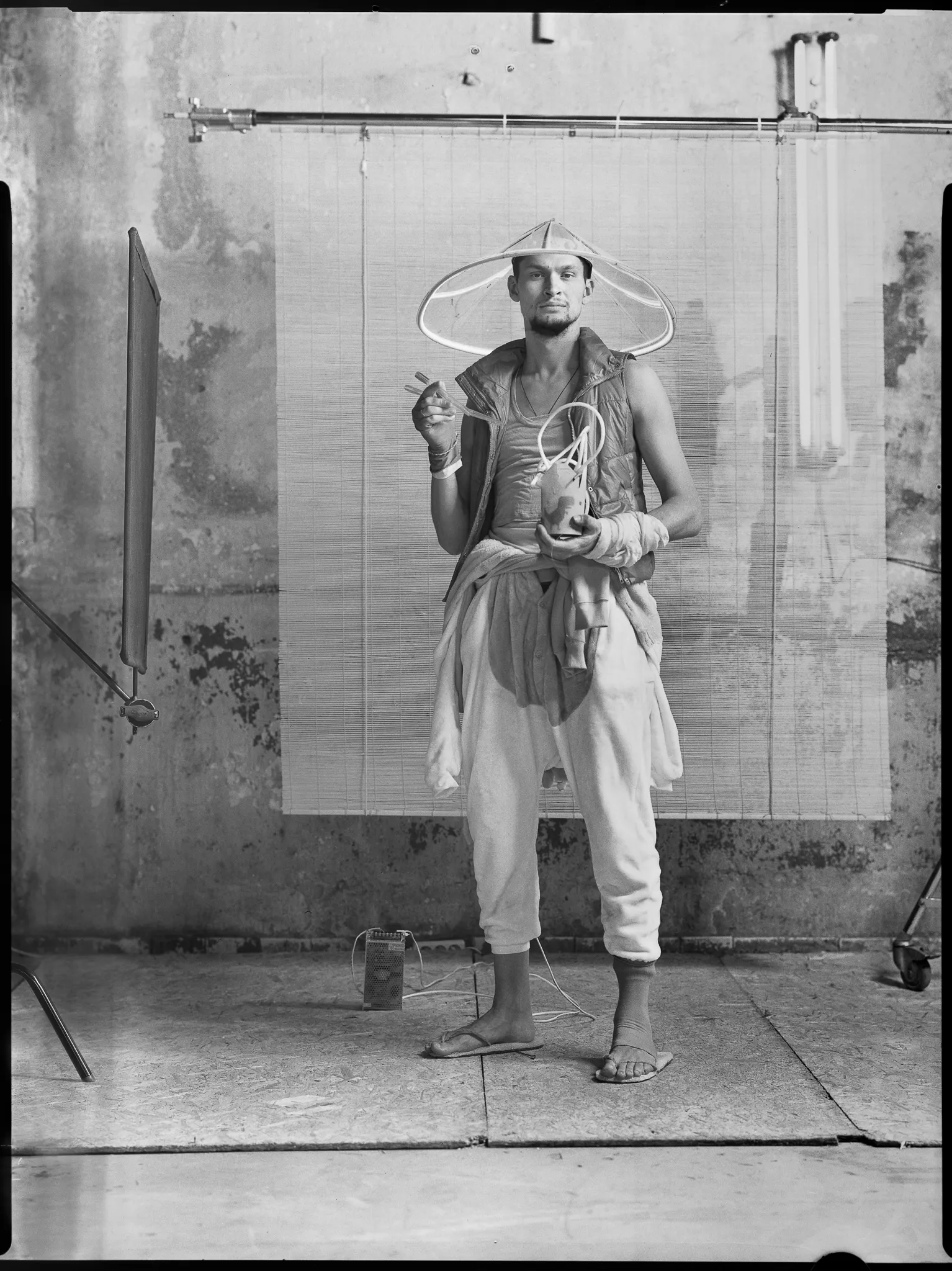 The Noodle Maker in a wide conical hat holds chopsticks and a cup of tube noodles, on black-and-white sheet film.