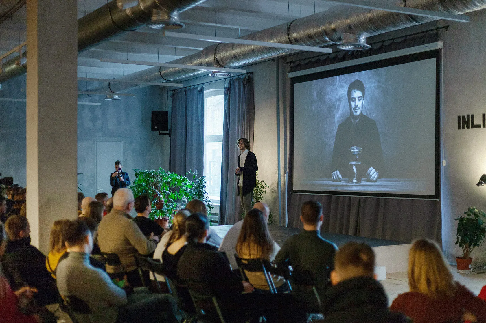 A speaker gives a talk to a packed loft hall, a black-and-white portrait on the screen beside him.