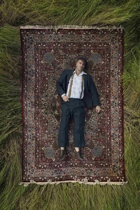 A man in a dark suit and braces lies on a patterned rug in long grass, seen from above.