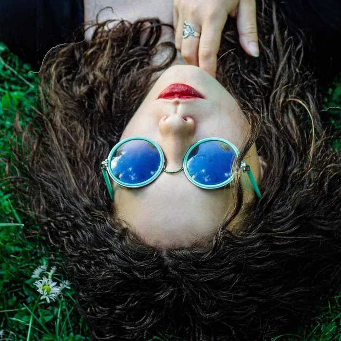 A woman with red lips and round blue sunglasses lies upside down in the grass.