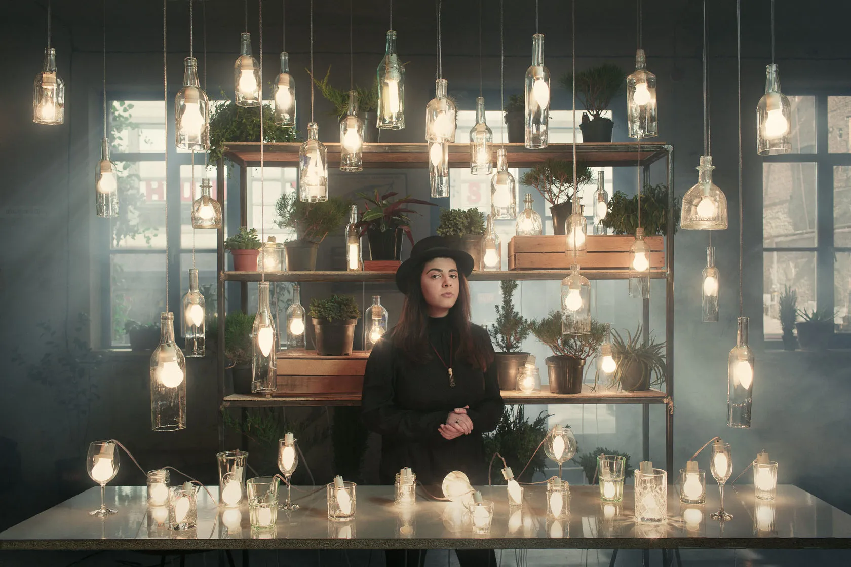Nina Naskidashvili in a black hat stands at a table of candles under rows of glowing hanging bottles.