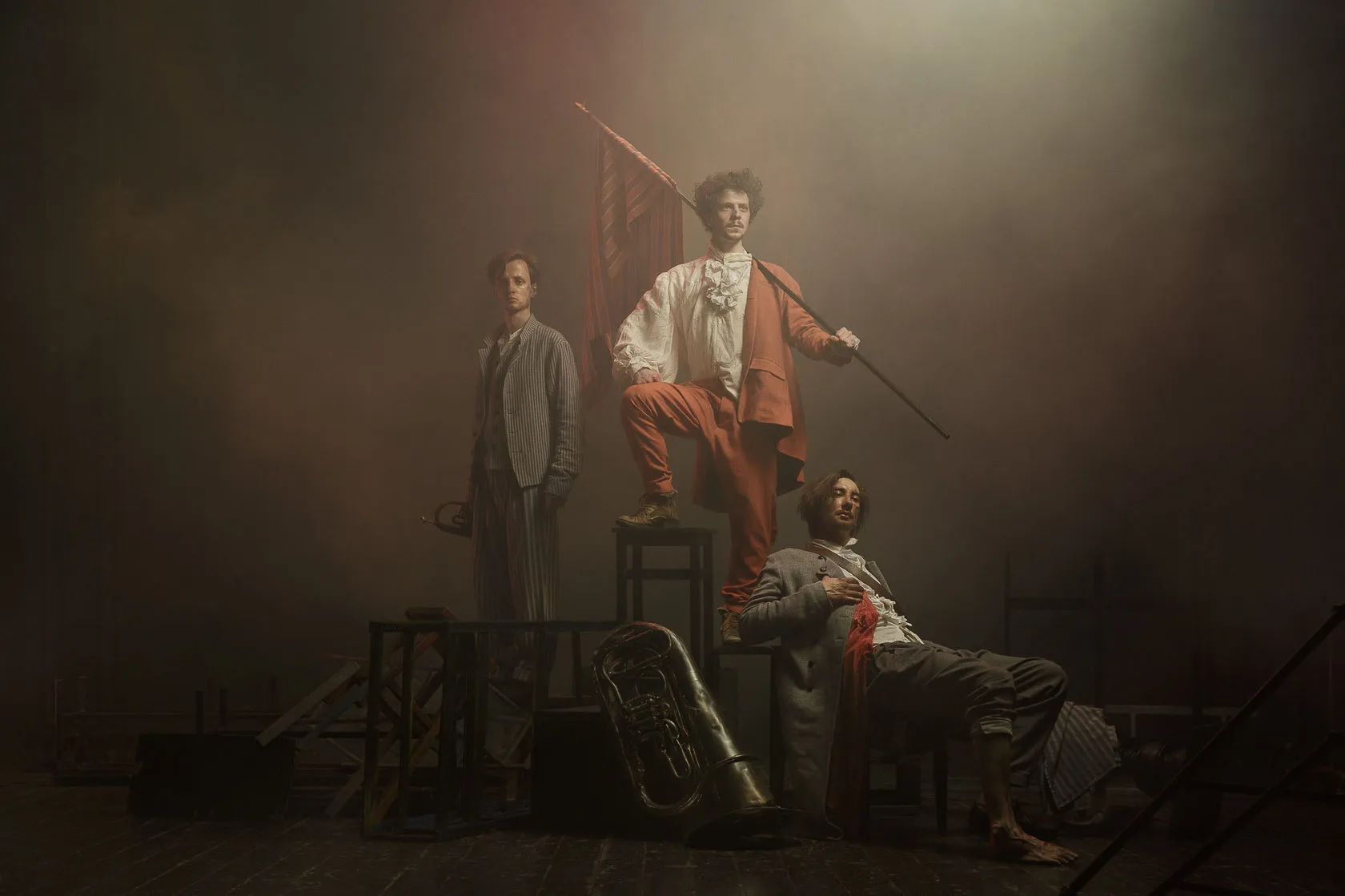 Three young actors in French Revolution costume pose on stacked chairs in smoke, one raising a flag.
