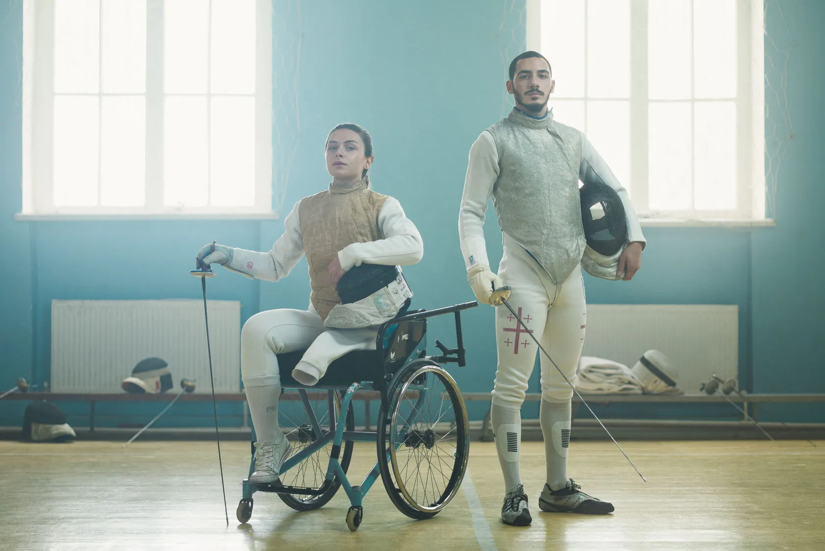 Two fencers, one in a wheelchair, pose with masks and blades in a sunlit gym.