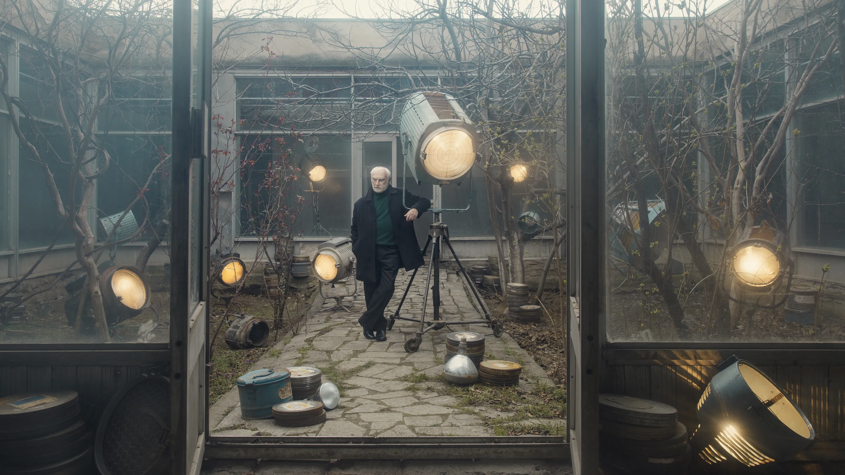 Online course by Andrei Kovalev: a grey-haired man leans on a film light in a glass courtyard, among old lamps, film cans and bare trees.