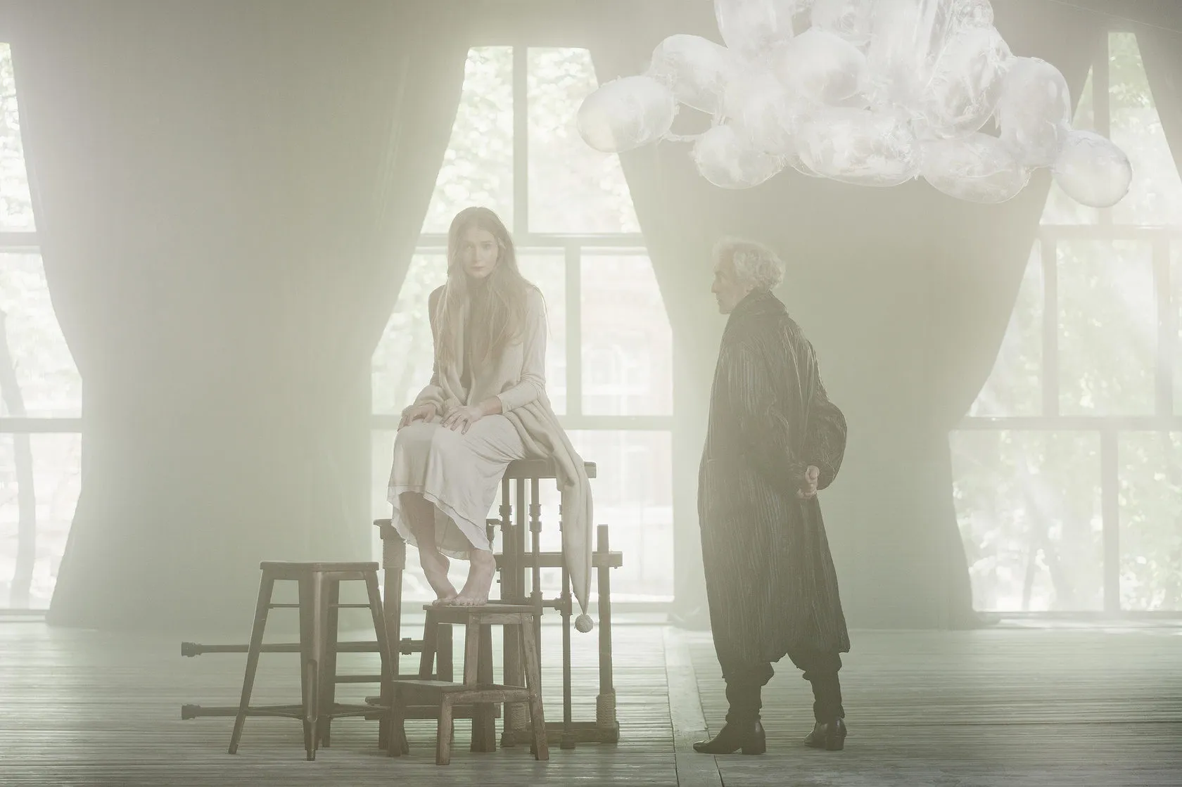 A young woman perches on stacked stools in a misty, curtained hall as an old man in a long coat looks on, a cloud hanging above.