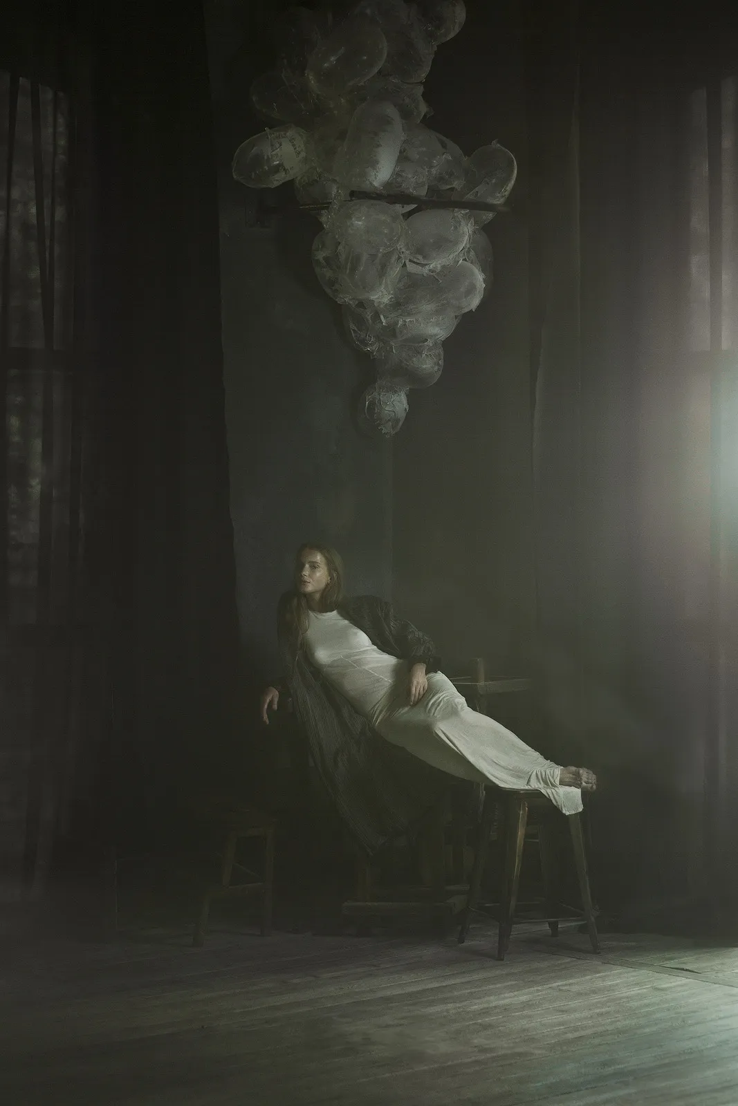 A woman in white lies back across stools in a dark hall, under a hanging cloud.