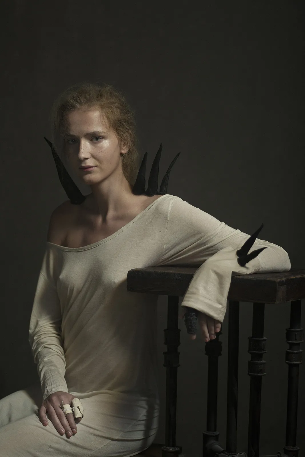 A red-haired woman in a loose white top leans on a dark banister, black feathers at her shoulder.