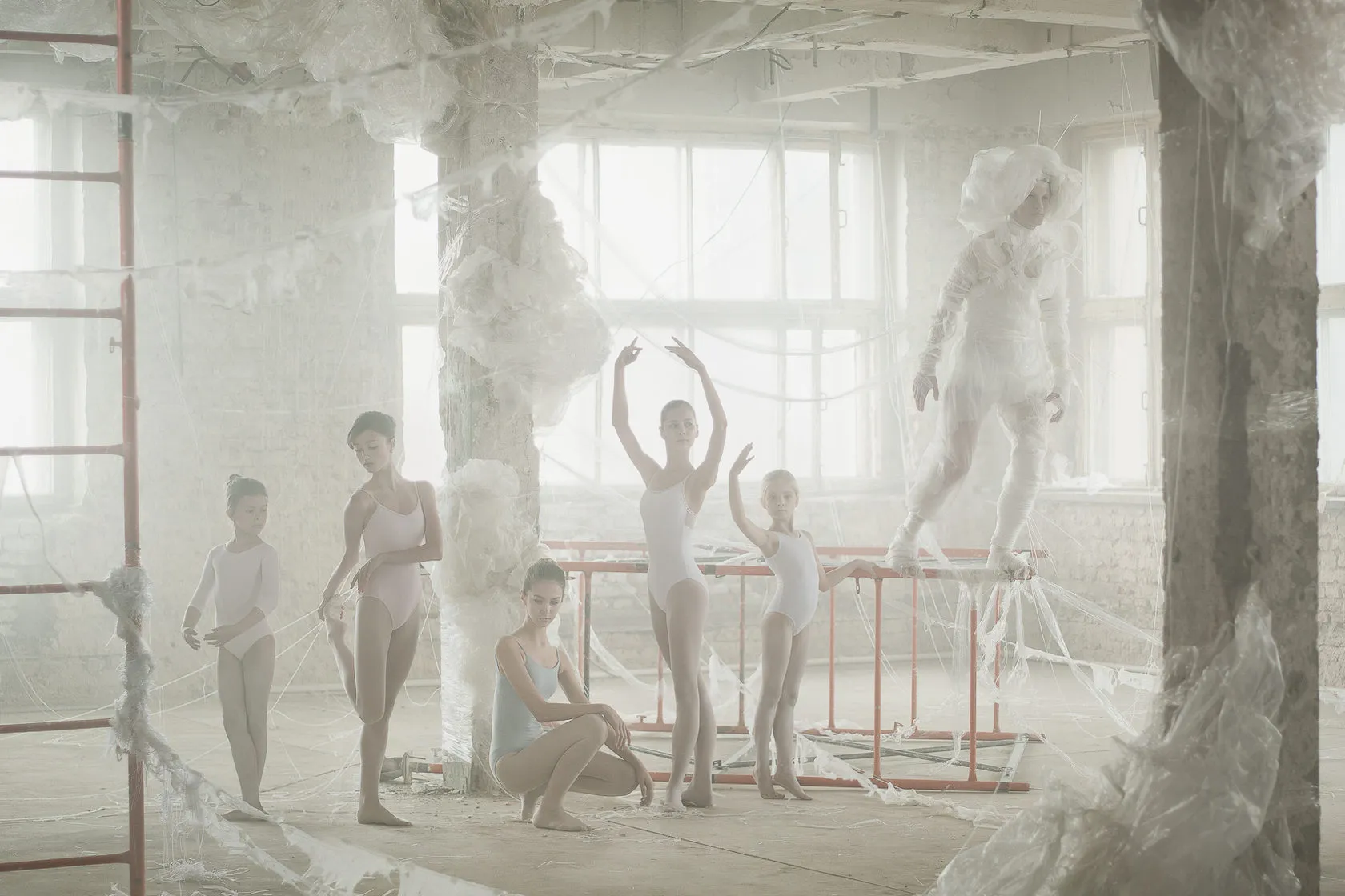 Young ballet students pose at the barres of a hall strung with white threads, a pale spirit behind them.