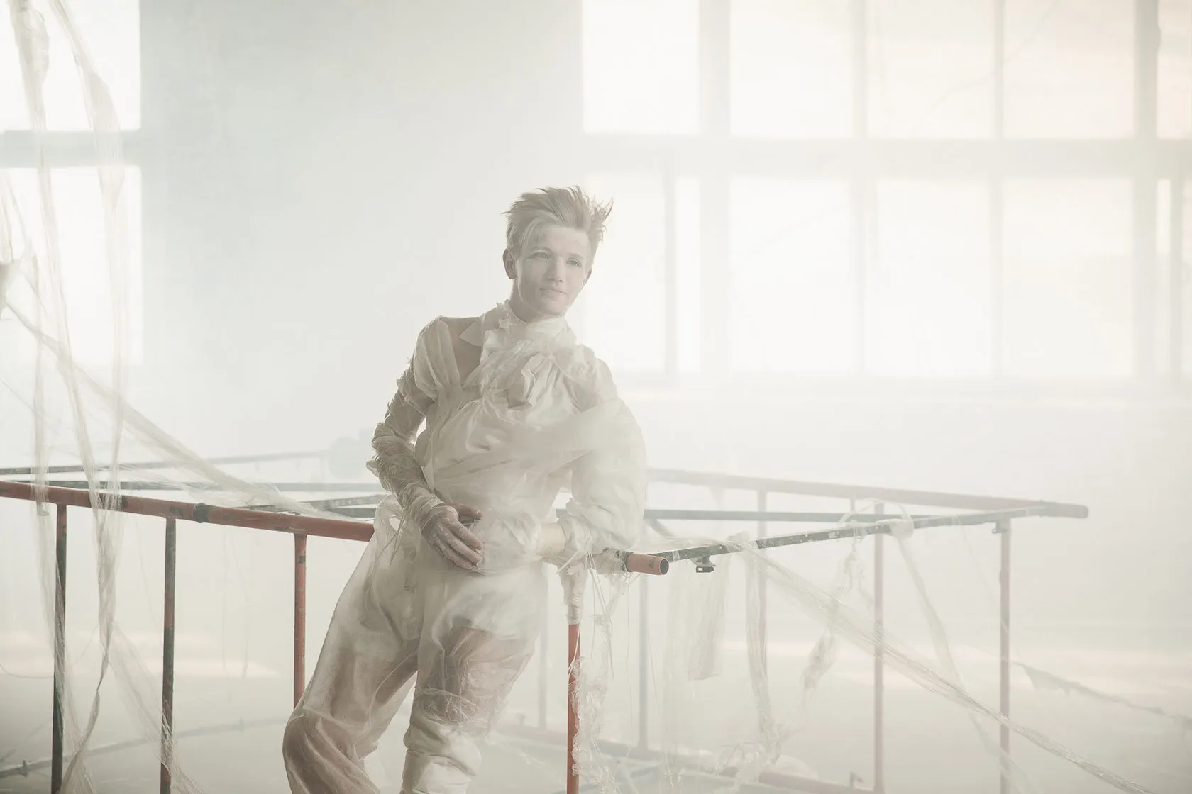 The spirit, played by Artemy Fogel, leans on a barre in a pale wrapped costume in a misty hall.