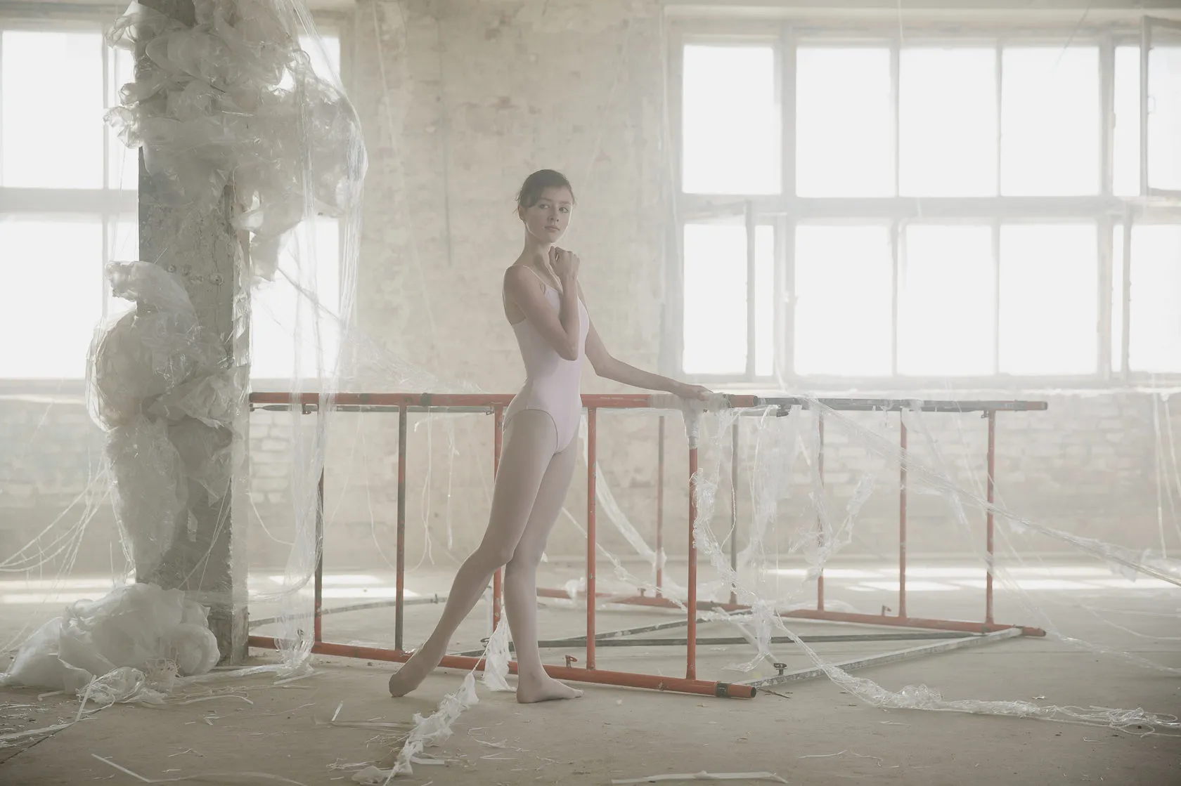 A ballerina in a pink leotard stands at a barre beside a column wrapped in white threads.