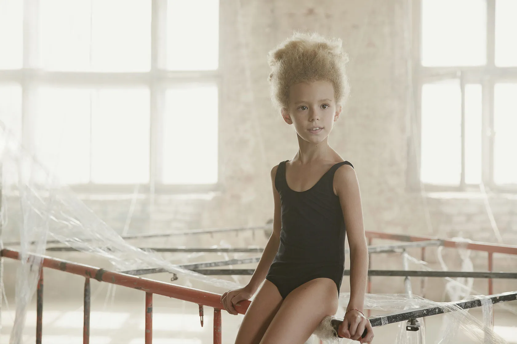 A young girl in a black leotard sits on a barre, her hair in a high bun.
