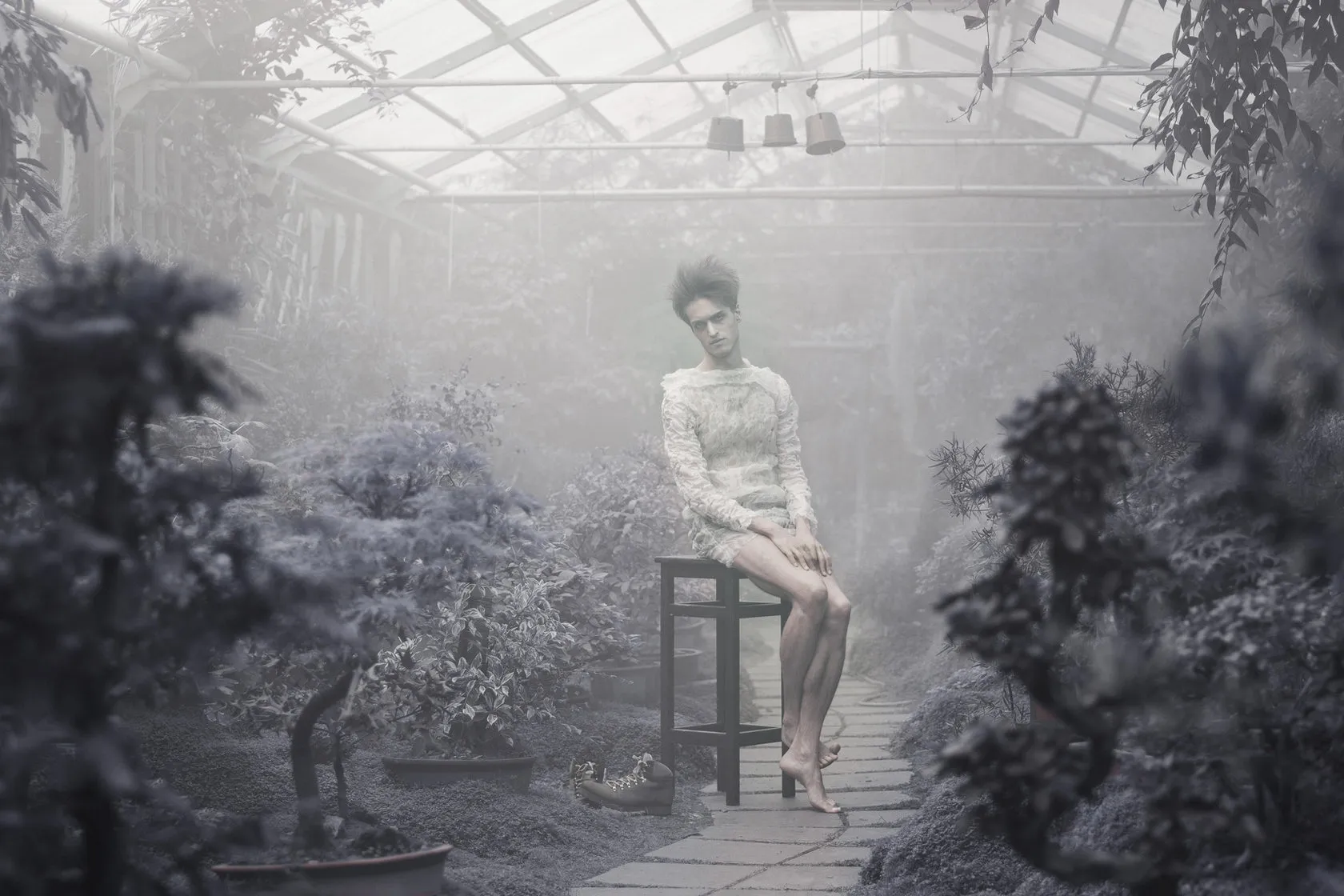 A woman in a lace dress sits on a stool in a misty greenhouse.