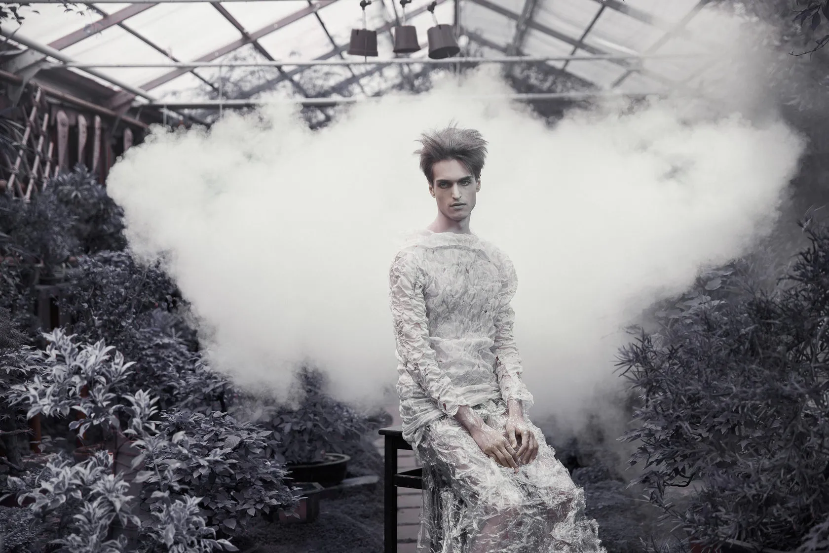 A young man in a lace suit sits in a cloud of white smoke among greenhouse plants.