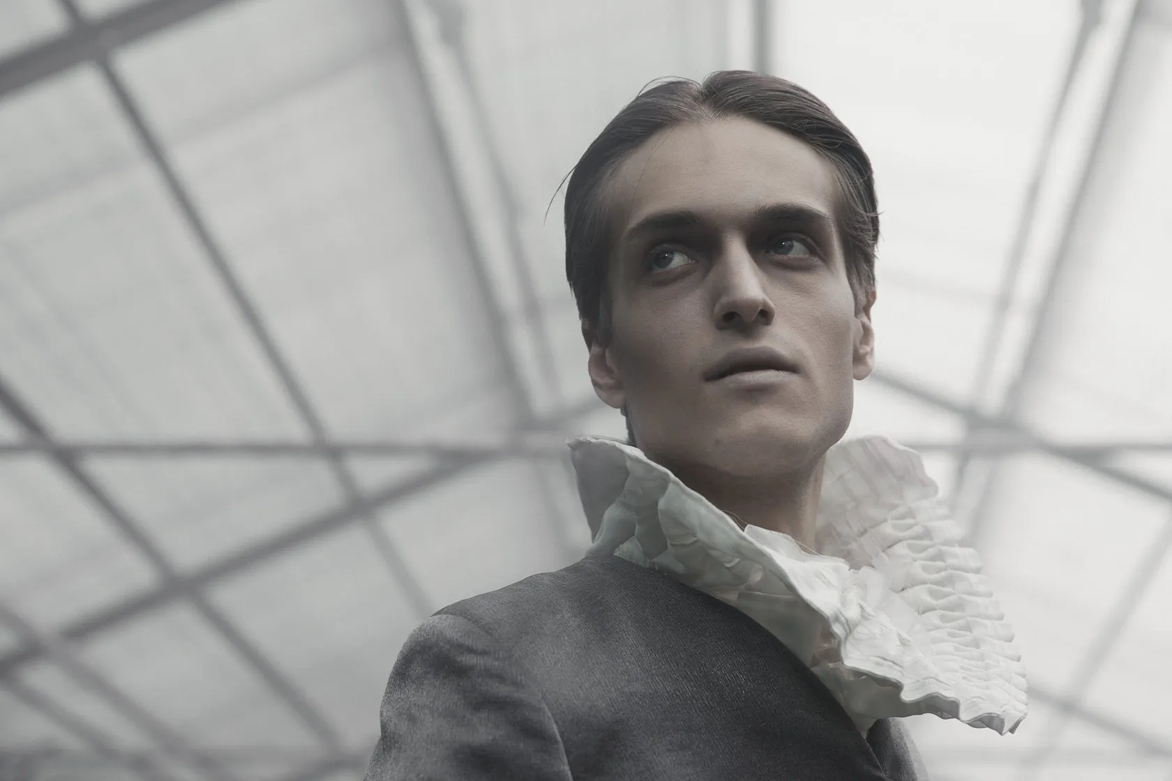 A young man in a ruffled collar under the glass roof of the greenhouse, close up.