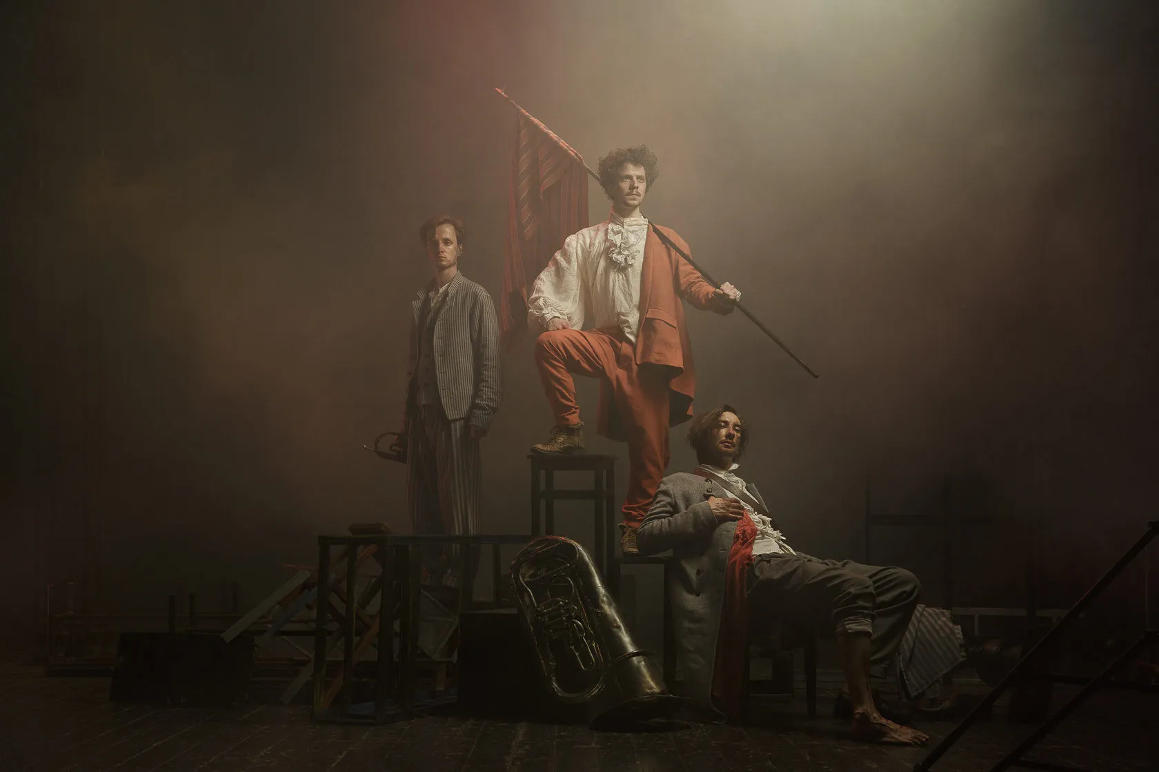 Three young actors in French Revolution costume pose on stacked chairs in smoke, one raising a flag.