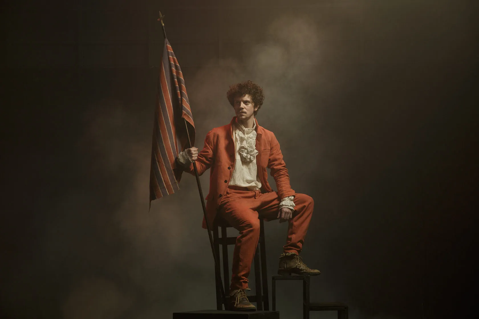 An actor in an orange suit sits high on a chair in drifting smoke, holding a striped flag.