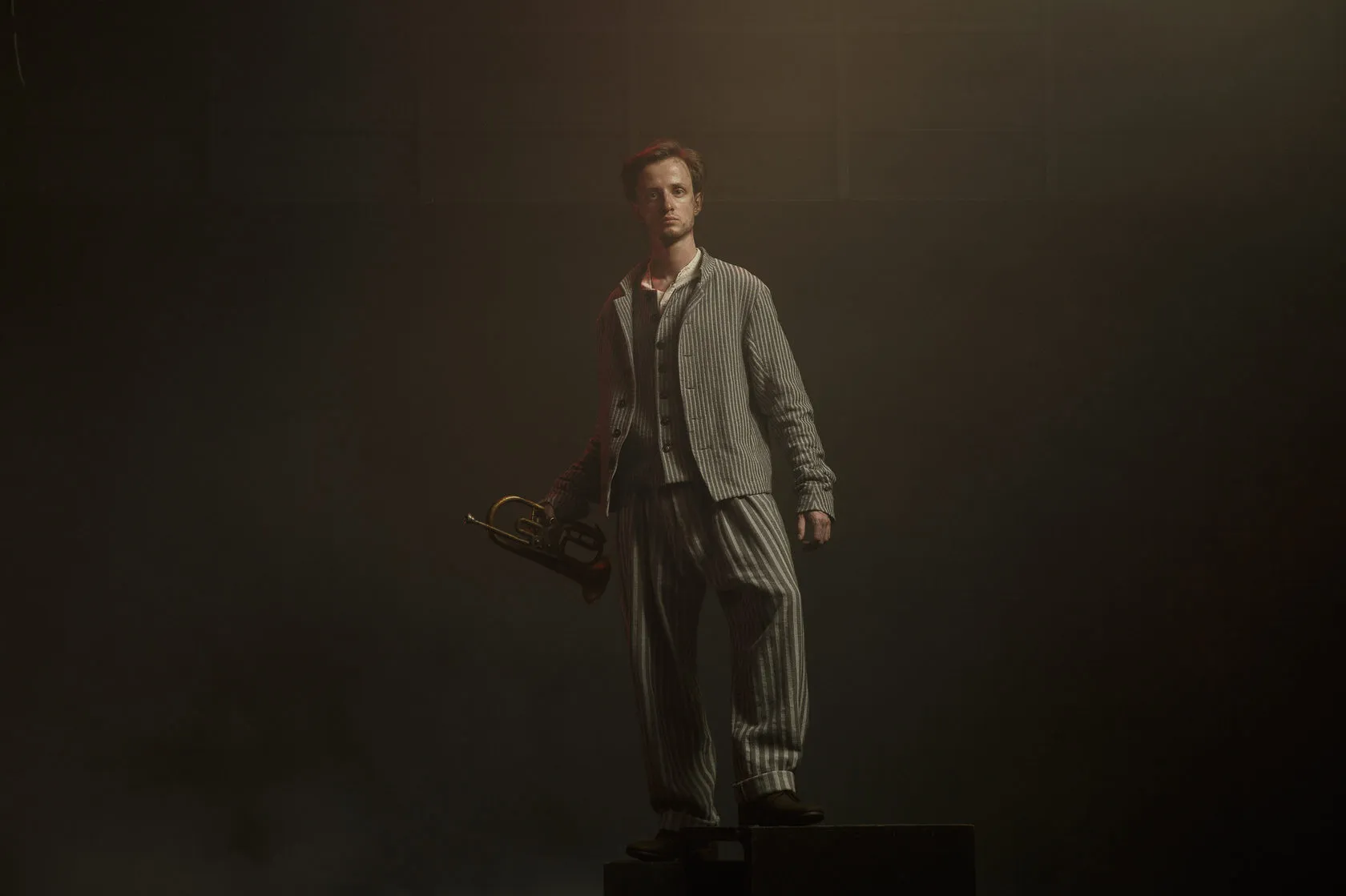 An actor in a striped suit stands on a dark box in smoky light, a brass horn in his hand.