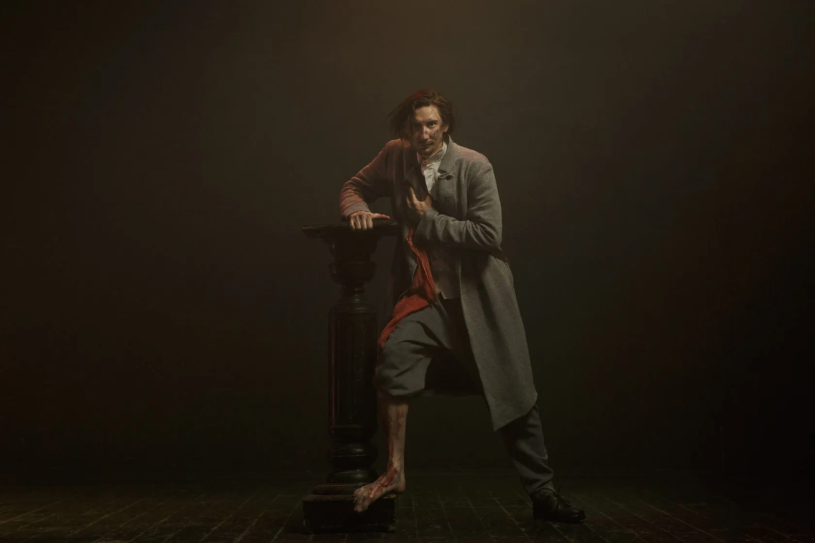 An actor in a long grey coat leans on a dark pedestal, one bare foot up on its base.