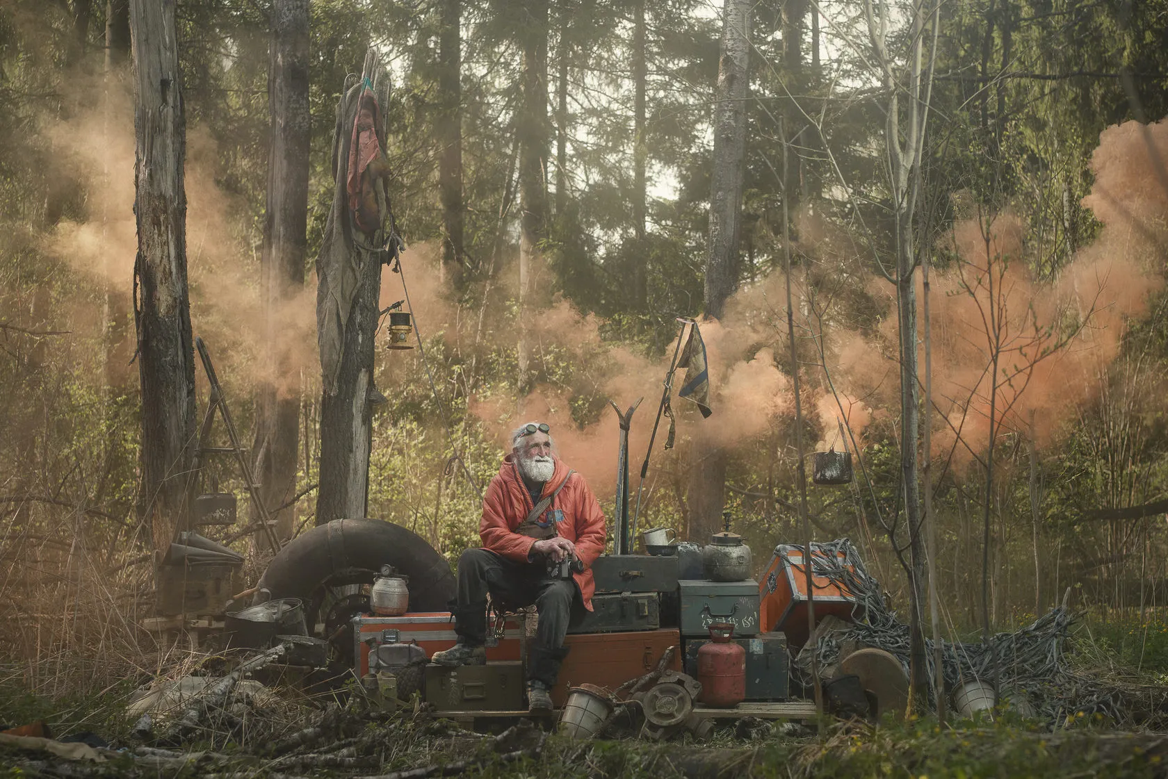 Vladimir Chukov sits on old luggage in a forest clearing as orange smoke drifts through the trees.