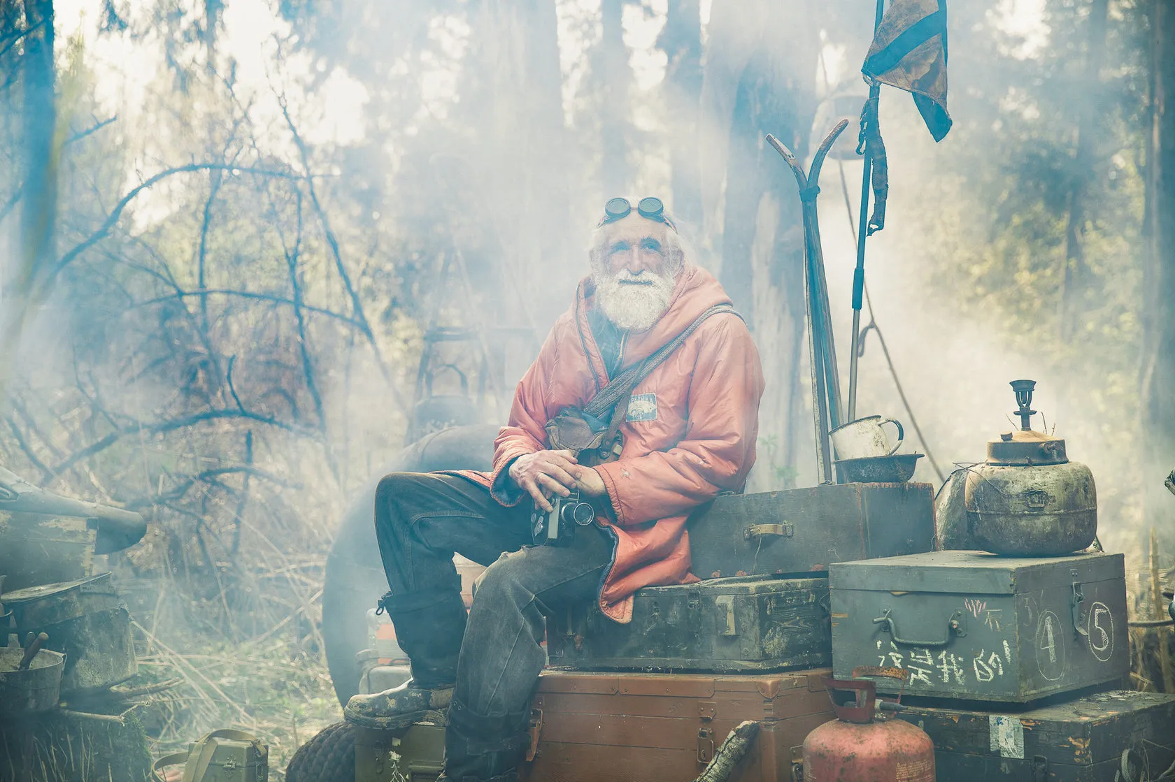 Vladimir Chukov in a red parka sits on old trunks in a misty forest, a kettle beside him.