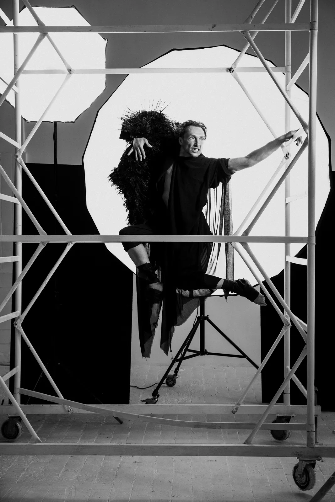 The actor in a black feathered cape perches on a scaffold in front of a large white light.