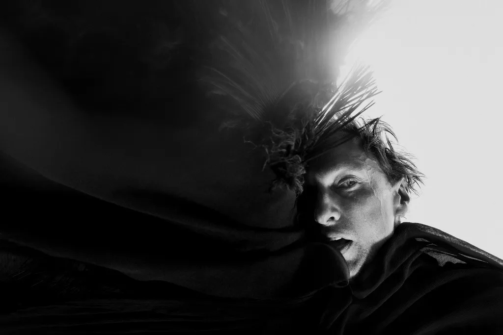 The actor in a black feathered headdress lies among dark folds of fabric, lit from above.