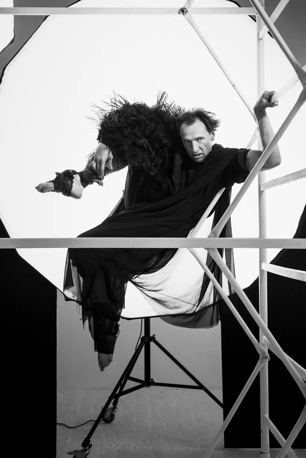 The actor in black feathers crouches on a scaffold, one leg stretched out, before a white light.