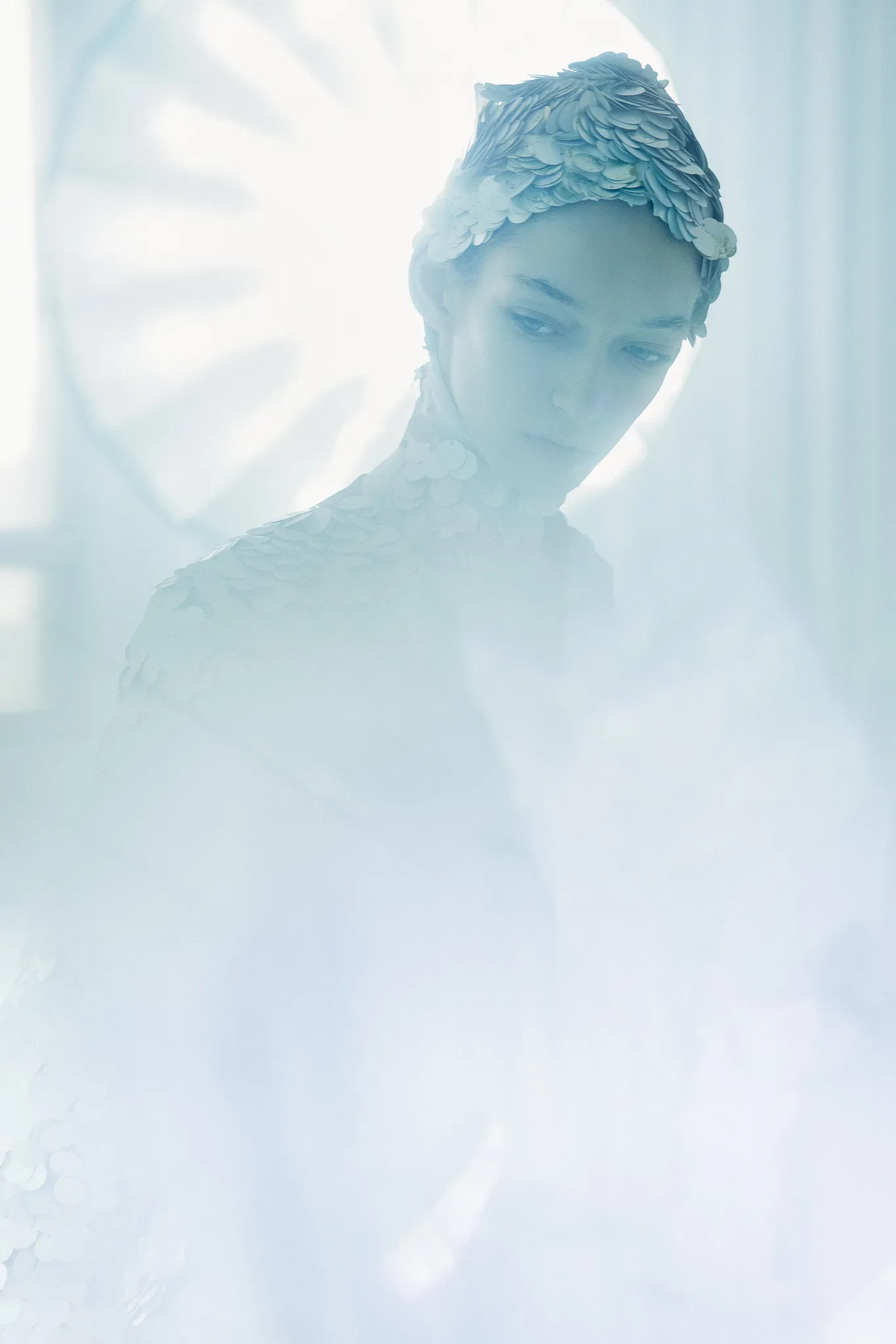 A model in a silver-scaled headpiece and costume turns away in soft blue light.