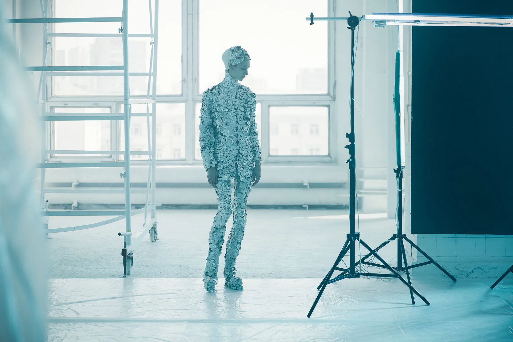 The model in silver scales stands in a bright white studio among scaffolding and light stands.