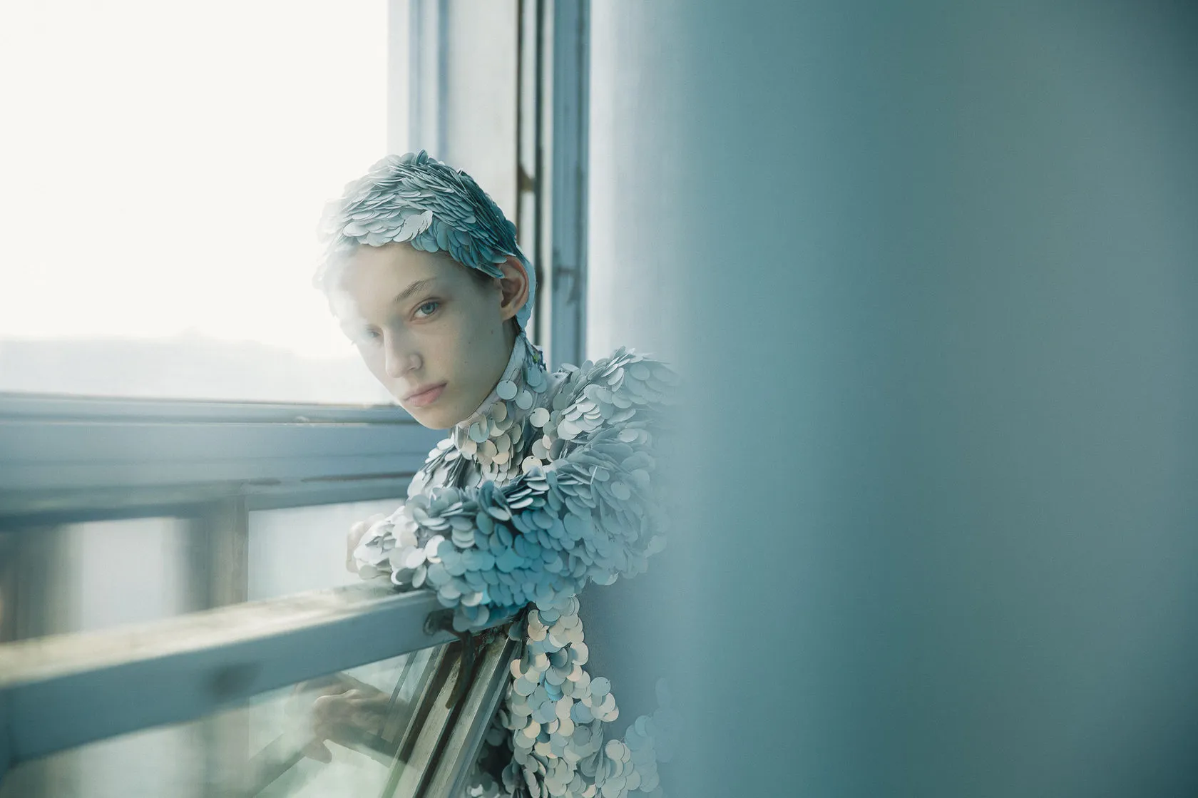 The model in silver scales leans on a windowsill and looks back over her shoulder.