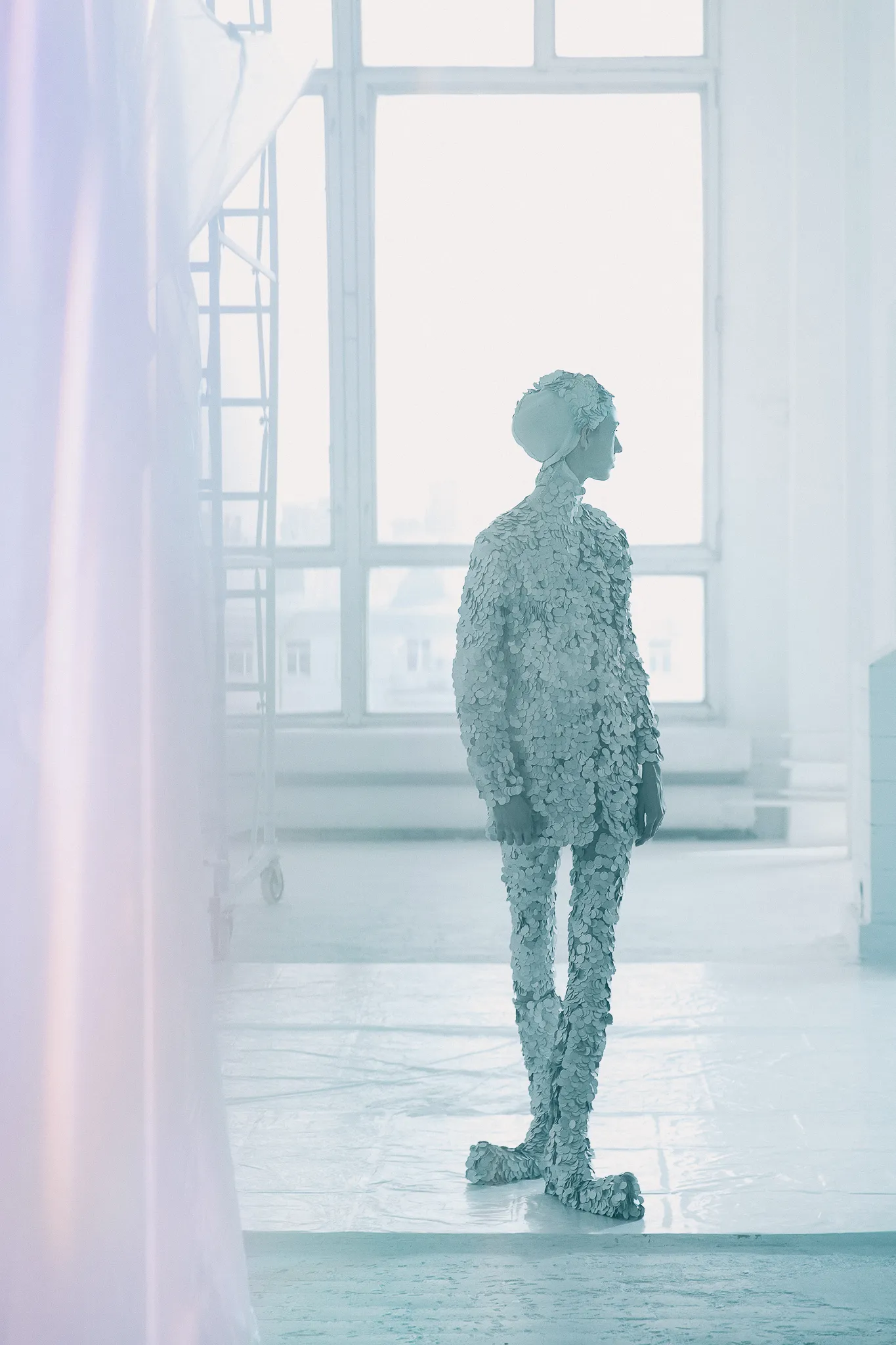The model in silver scales stands by a tall, bright studio window.