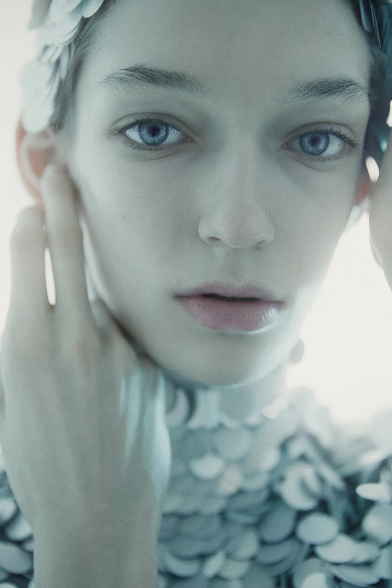 The model in close-up, her hands framing her face, silver scales at her neck.
